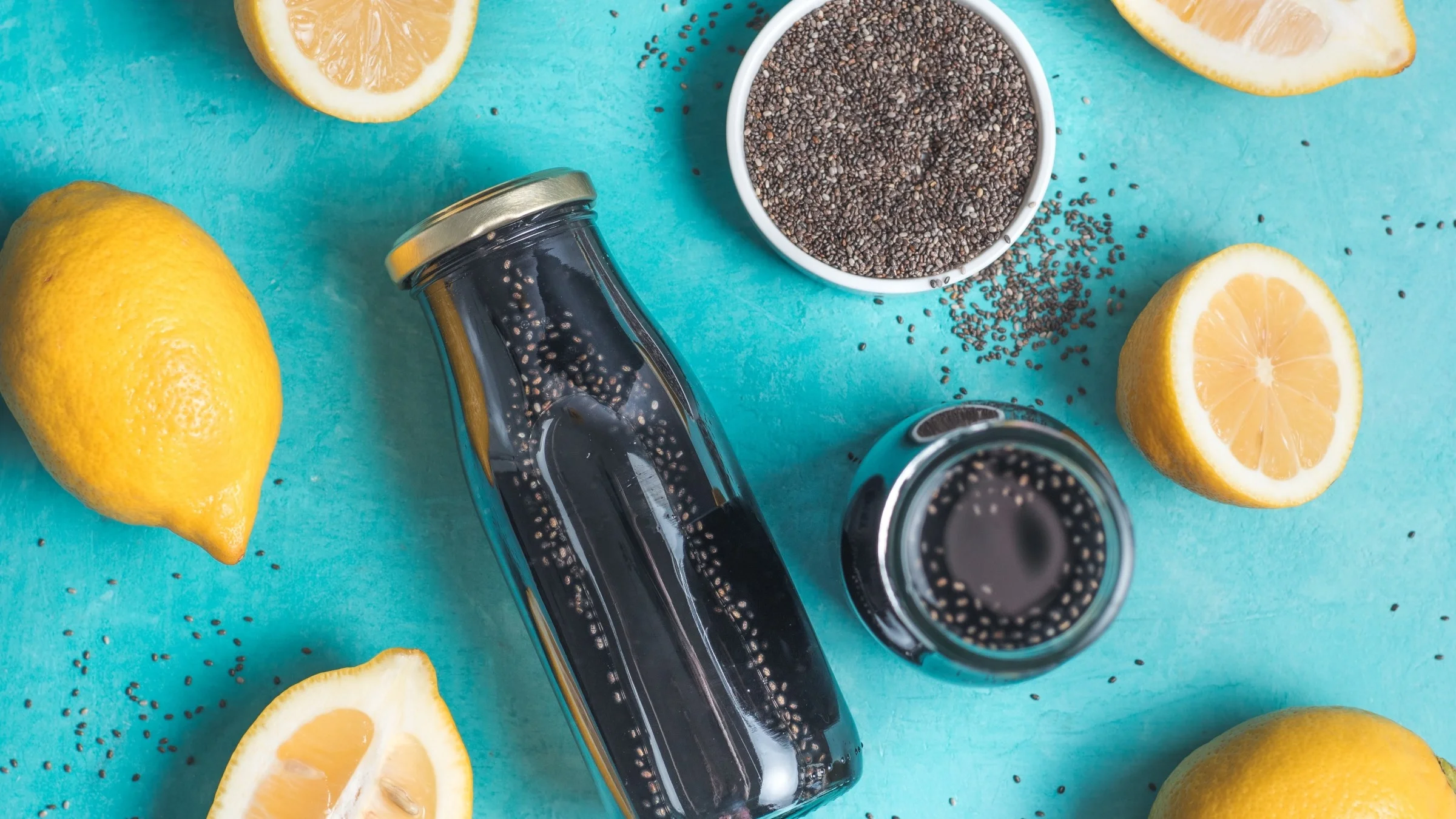 A glass bottle of activated charcoal detox mix laid flat on a teal background surrounded by lemons and chai seeds.