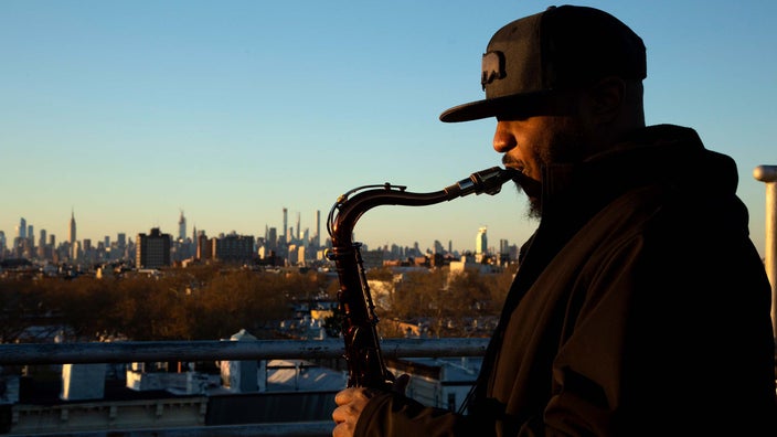 Side profile of James Casey playing his saxophone outside. The NYC cityscape is in the background.