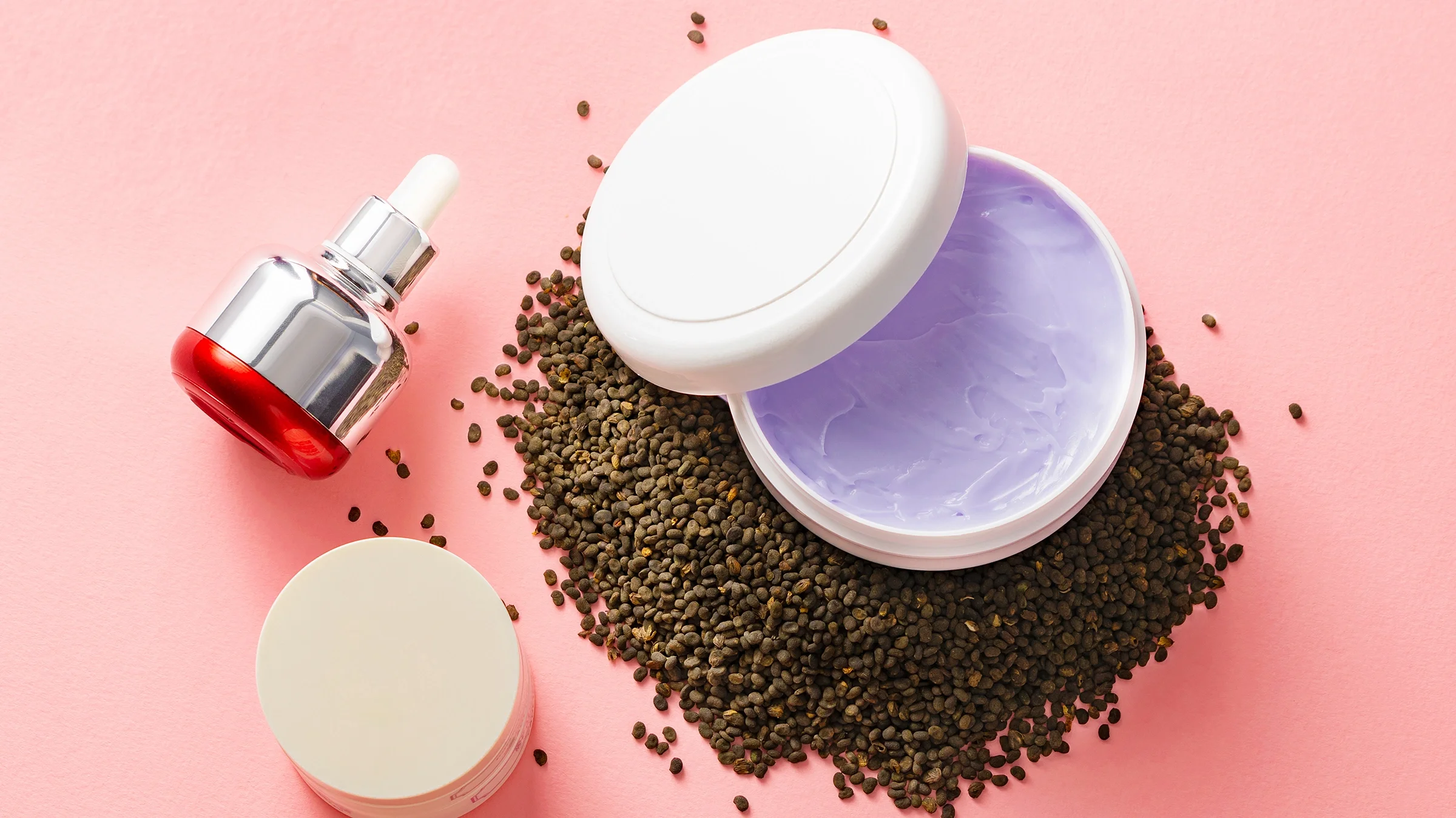 A still-life of cosmetic products with babchi seeds is shown on a pink surface.