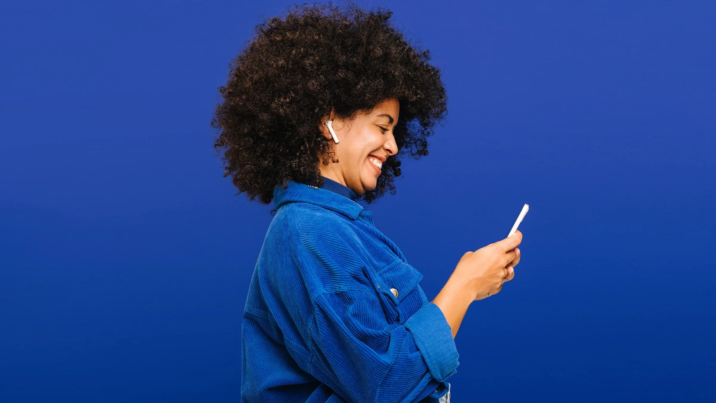Smiling woman, wearing earbuds, and looking down at a smartphone, against a blue background.
