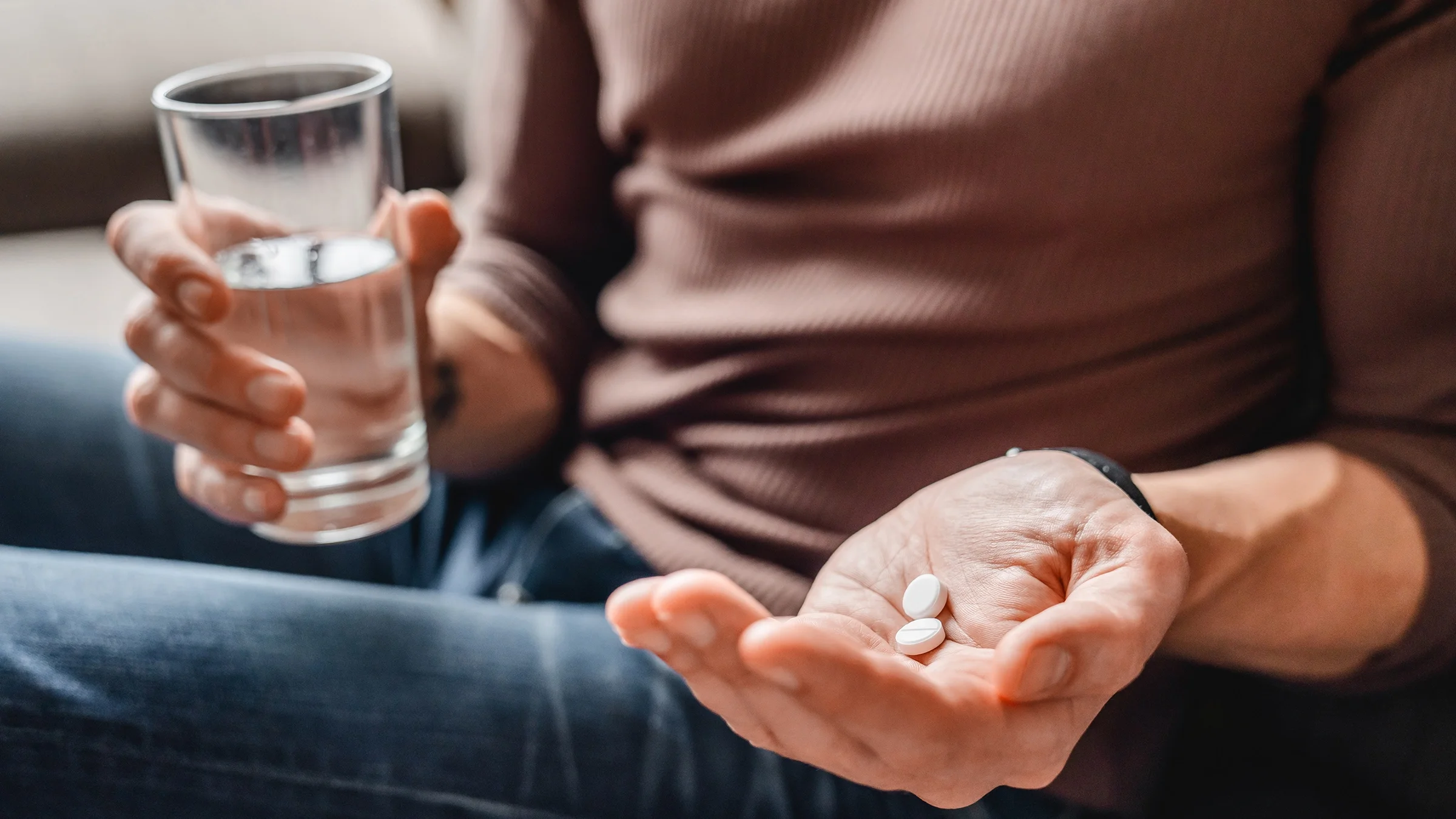 Cropped close-up of a man taking two white pills with a glass of water.