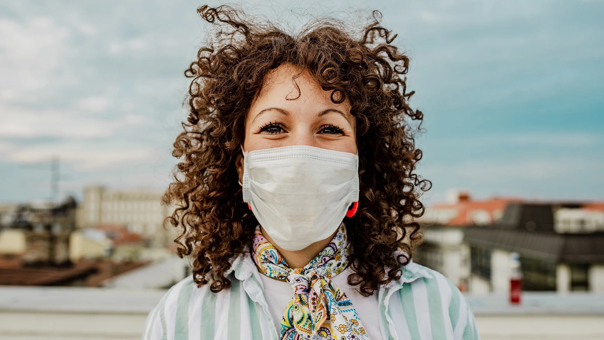Portrait of a young woman with curly hair blowing in the wind outside wearing a medical face mask smiling at camera