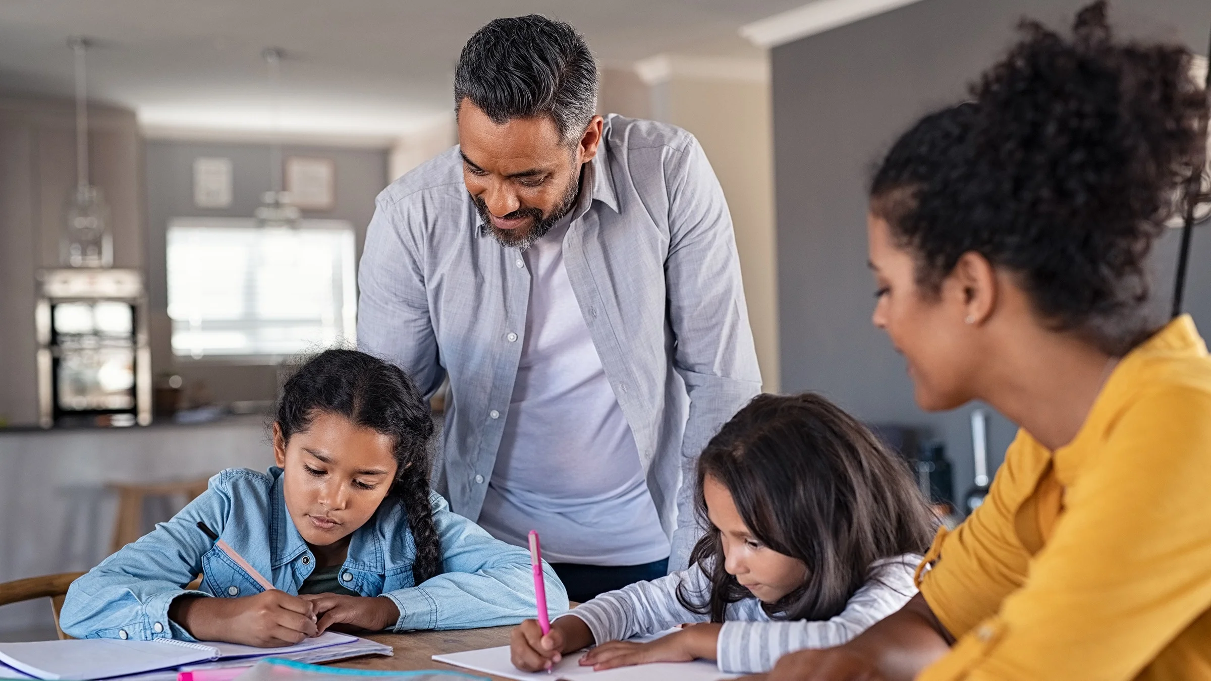 Family at the dining table doing homework. Both parents are standing and waiting by to help out while two children do their assignments.