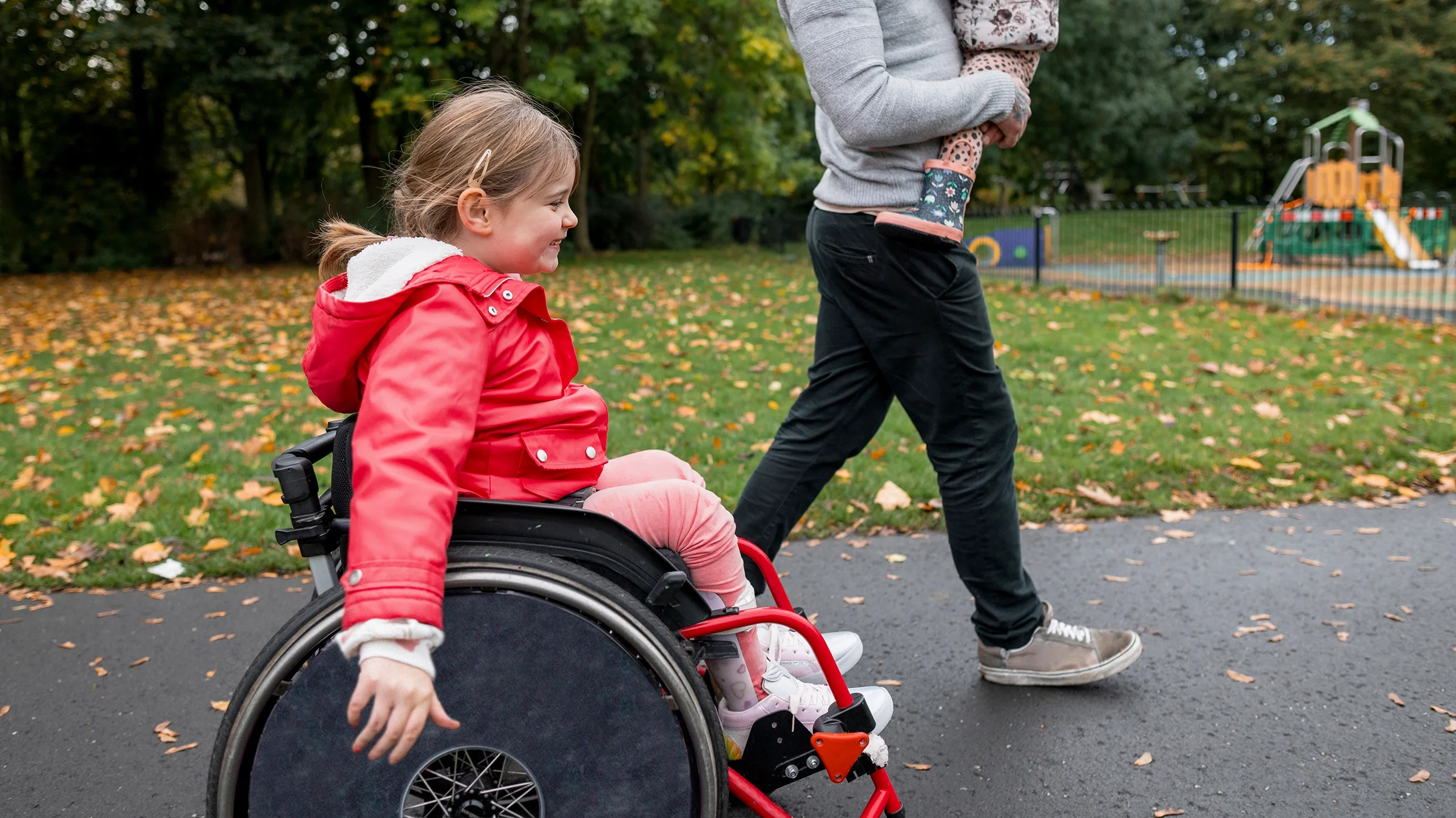 A young girl is in a wheelchair going through the park with her dad and sibling.