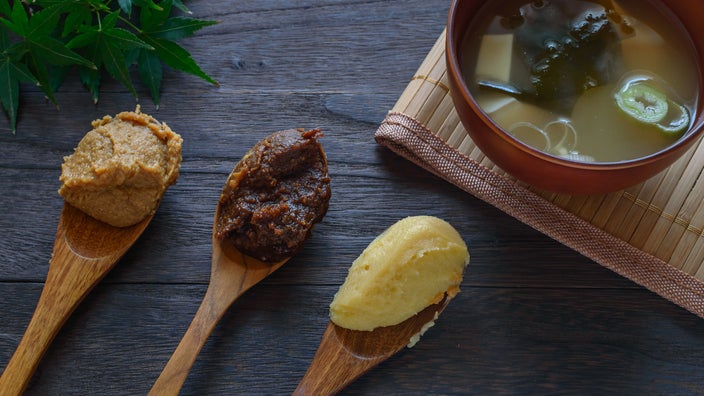 Three wooden spoons, each holding a different type of miso, on a tabletop next to a bowl of miso soup.