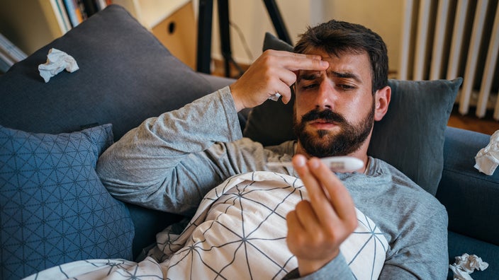 Man with beard holding his head and taking his temperature while sick at home.