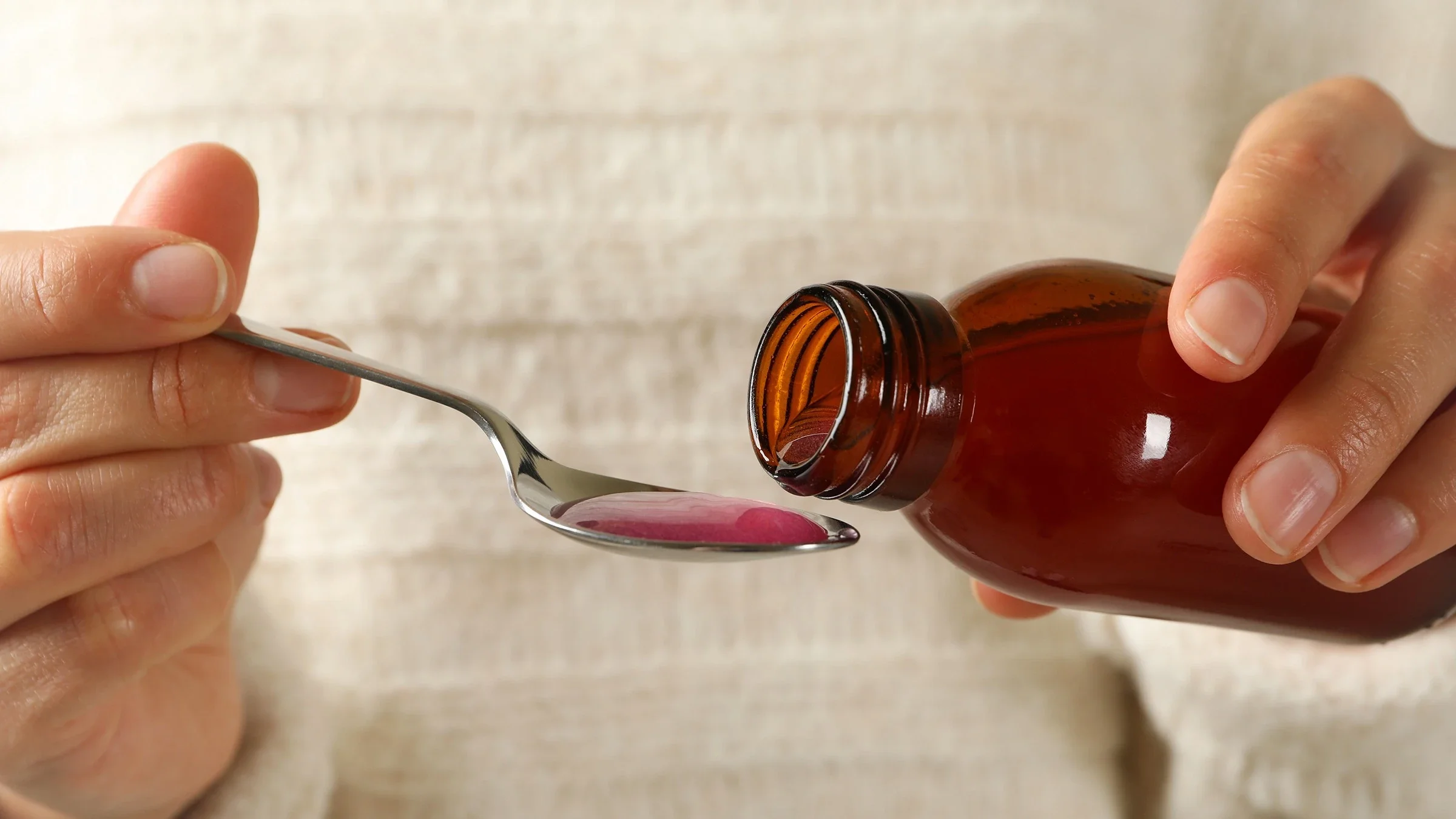Close-up of a person’s hands holding a liquid medicine bottle and spoon. There is already a full spoonful of pink liquid. The person is wearing a white textured sweater.