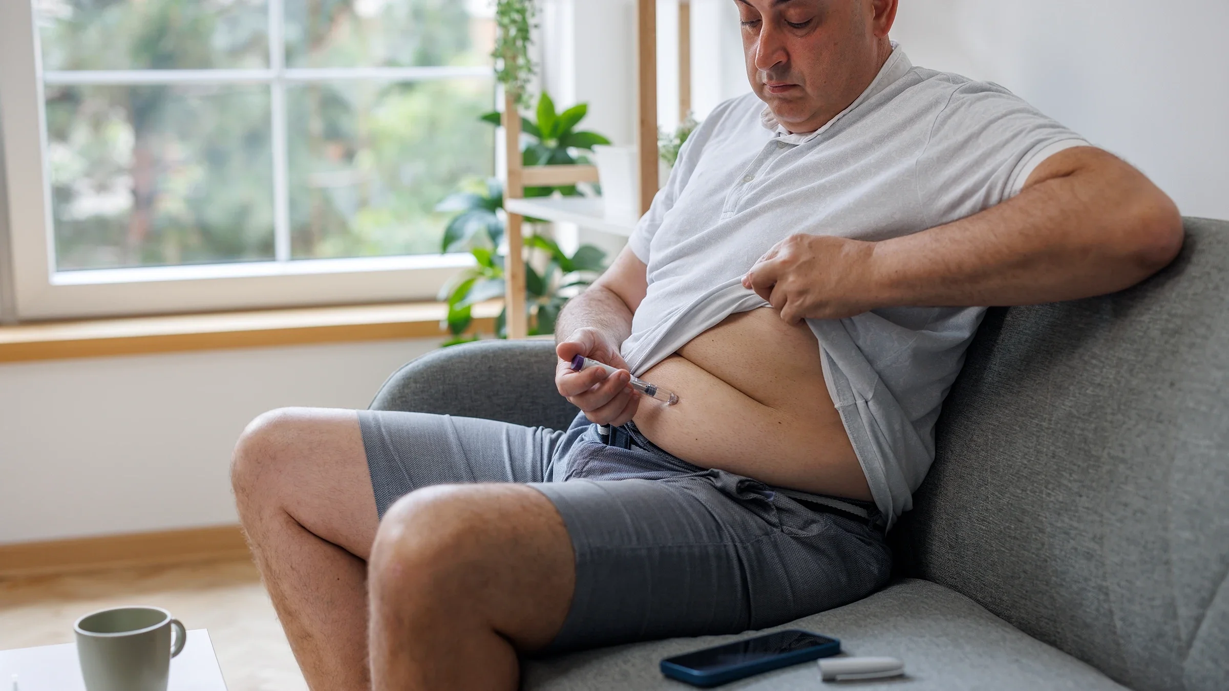 A man sitting on the sofa injects insulin in his abdomen.