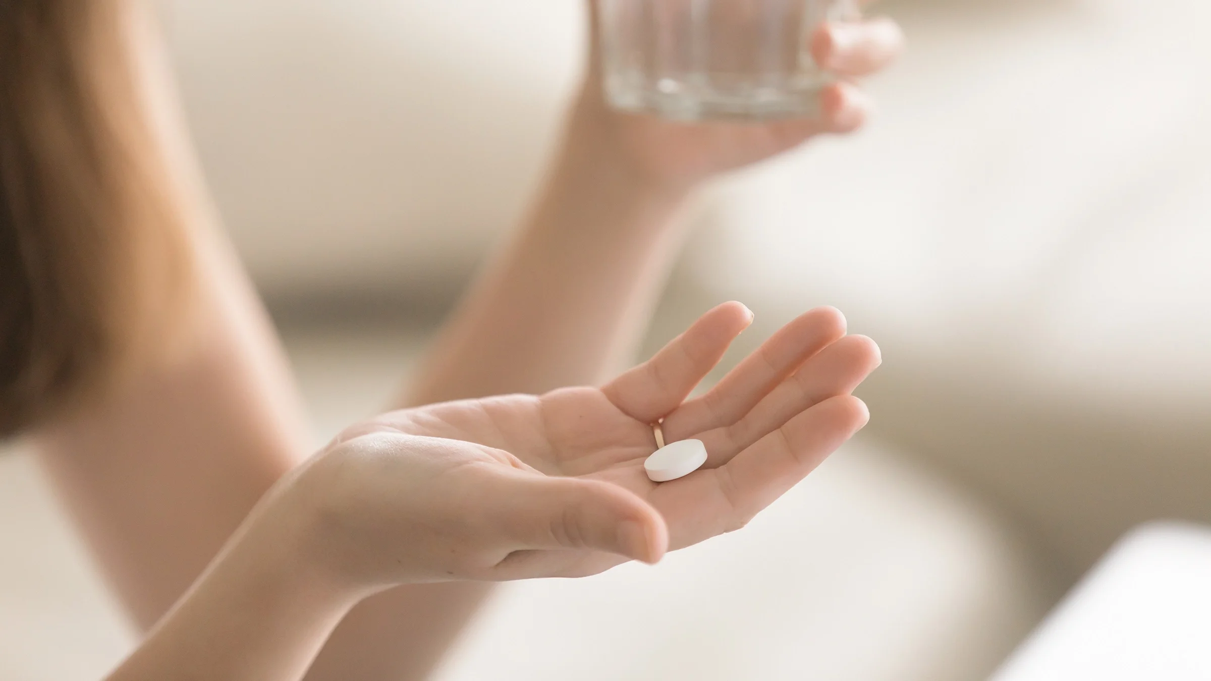 Cropped shot of a woman's hand holding a single white pill. In her other hand there is a glass of water.