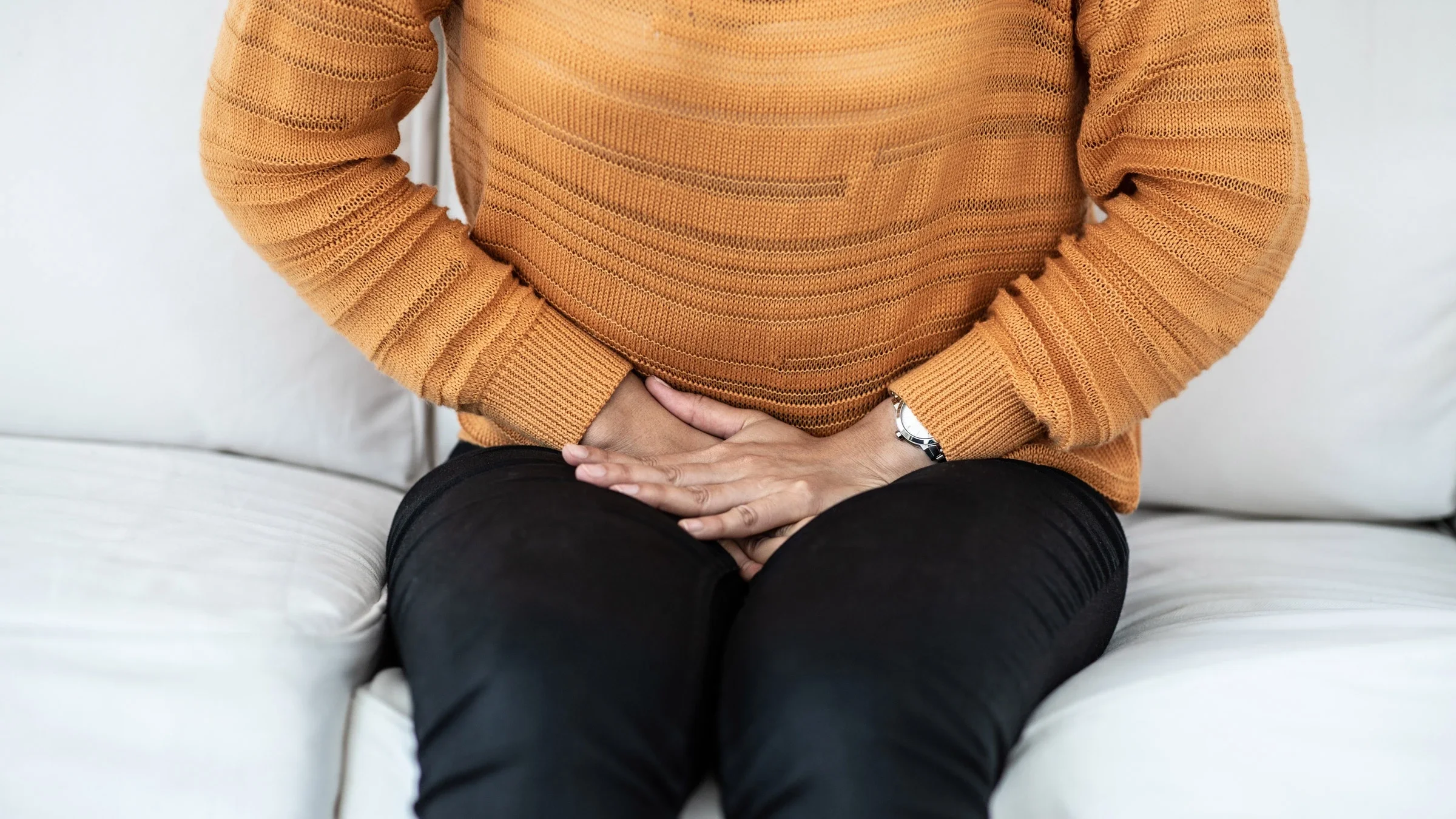 Close-up of a person wearing an orange sweater and black pants. They are pressing their hands into their pelvis in pain. 