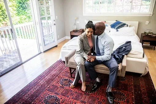 A mature Black man and an Indian woman cuddling up at foot of the bed while getting ready for the day.
Adam Kaz/E+ via Getty Images