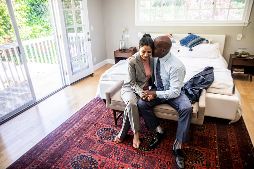A mature Black man and an Indian woman cuddling up at foot of the bed while getting ready for the day.