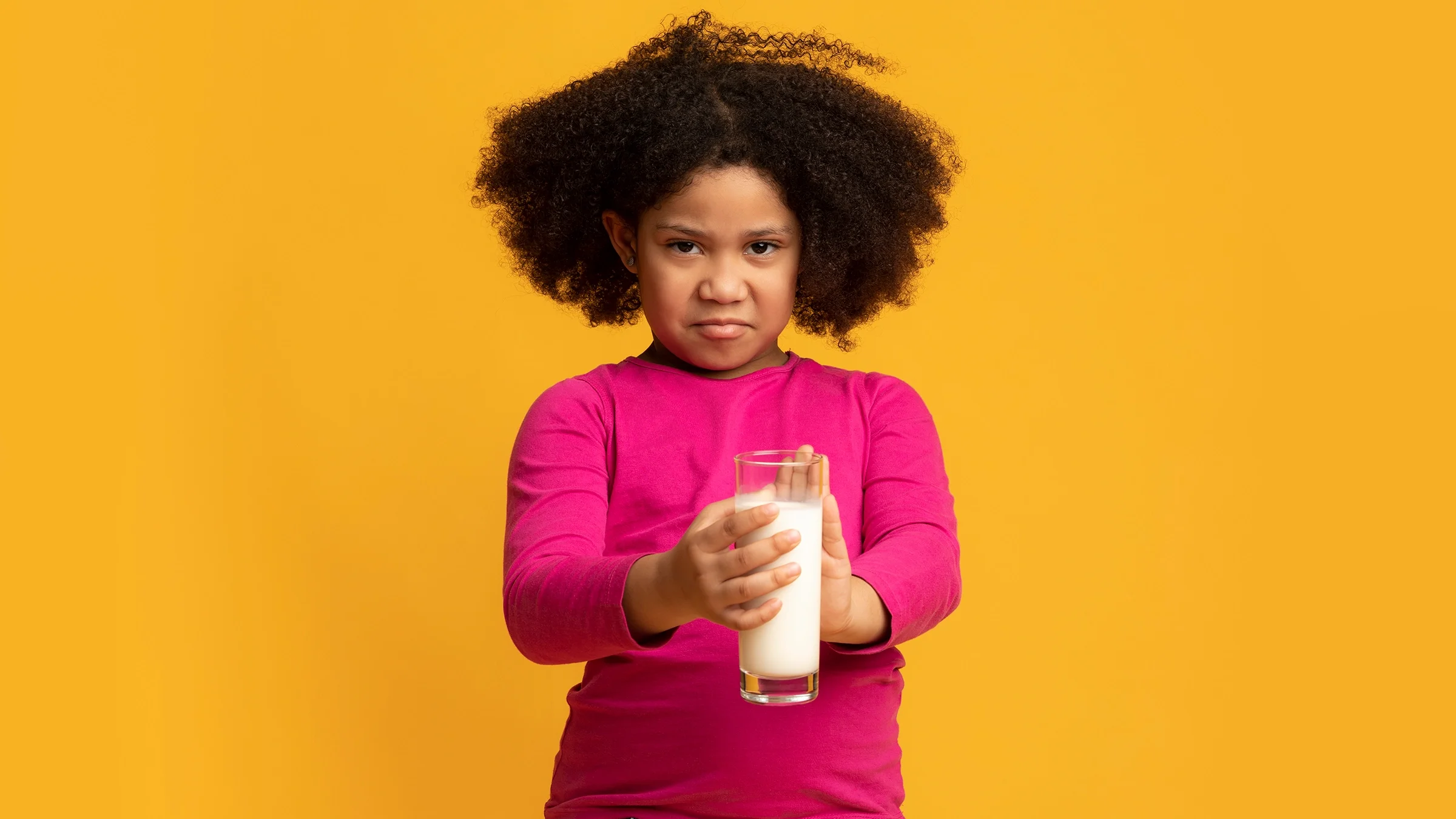 Young girl with curly hair with a look of disgust on her face. She is holding a glass of milk and on a yellow studio background.