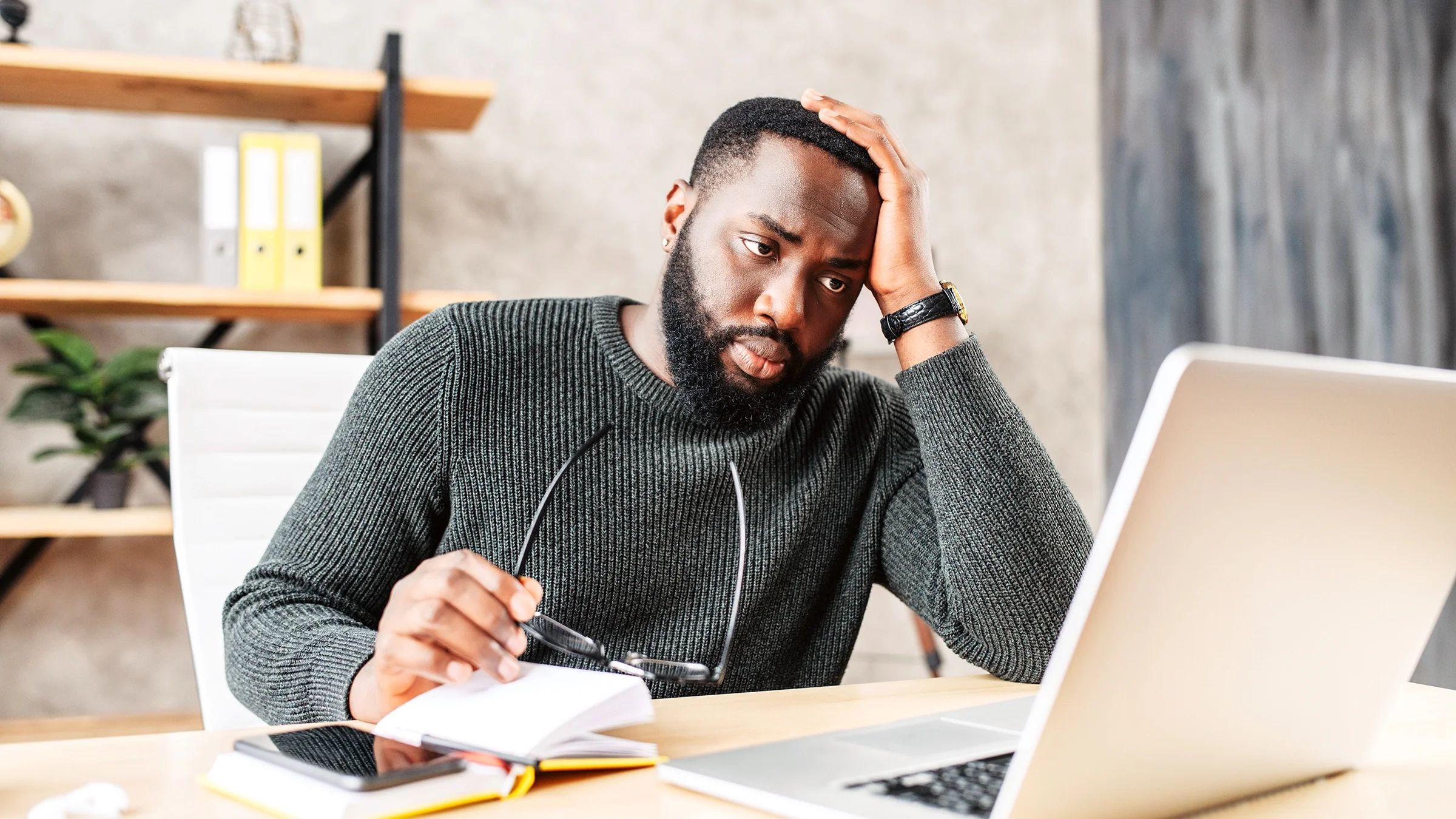 Man sitting at his laptop at home with his glasses in one hand and his other hand on his forehead in worry.