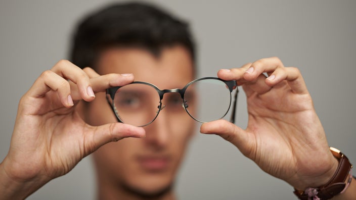 Young man with short black hair holding out his glasses to the camera. The glasses are in focus in the foreground while he is out of focus in the background.