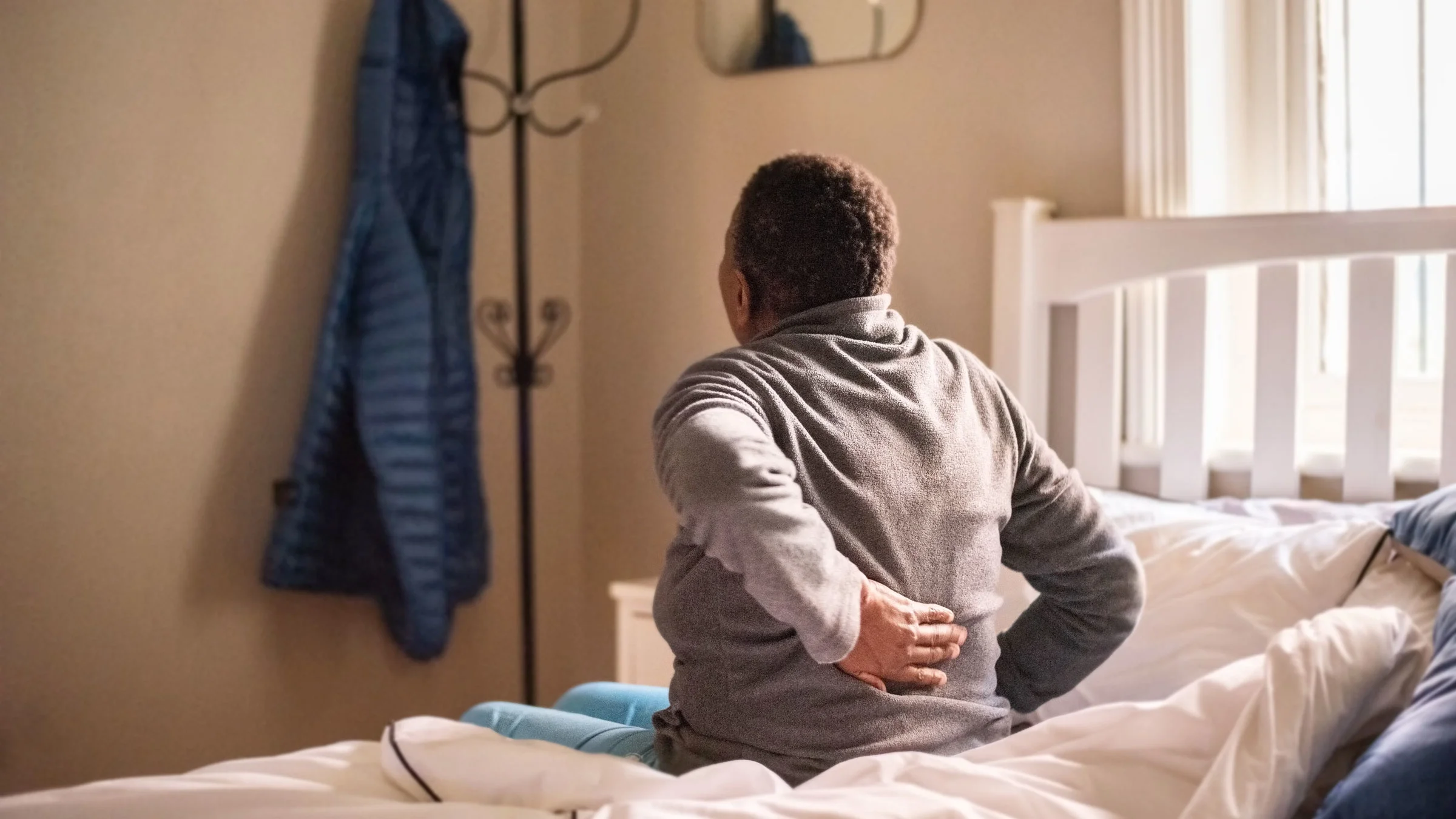Rear view of a senior woman sitting in her bed in the morning holding her back.