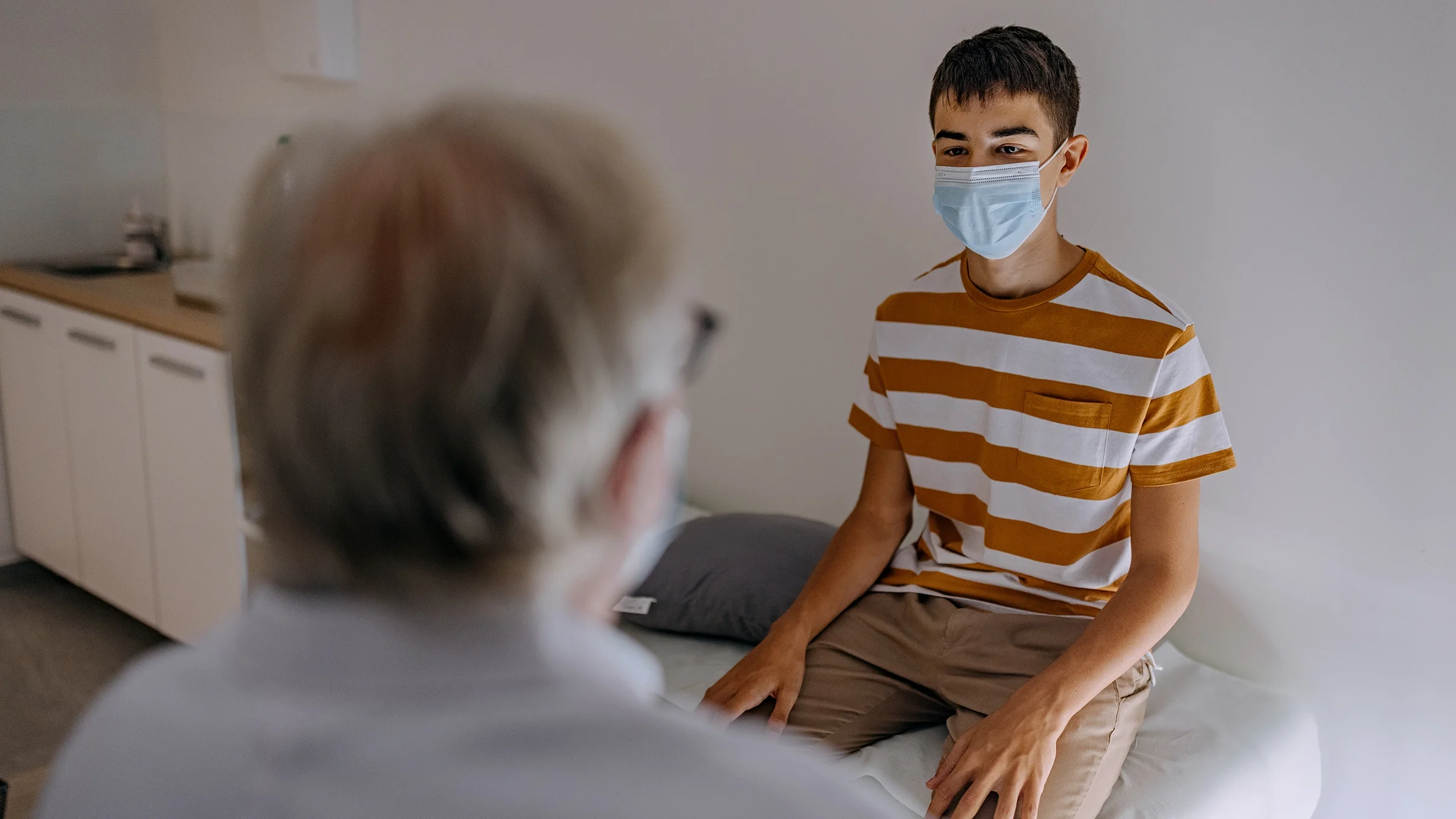 A teenager is in the exam room with a provider. The teen is wearing an orange and white shirt with stripes and a blue medical mask.