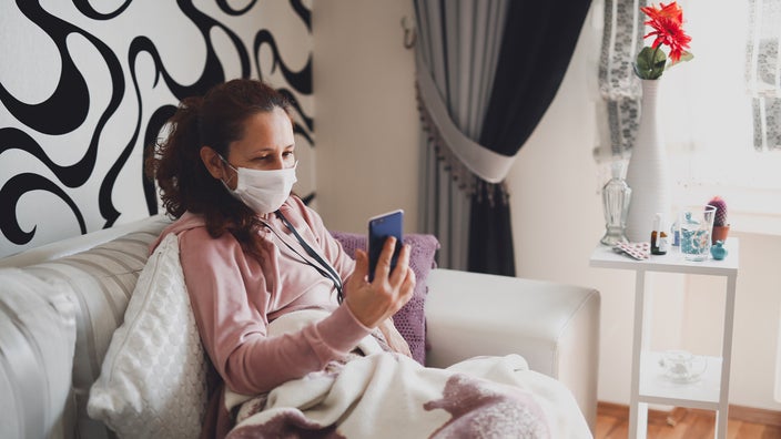 Middle aged woman sitting on her couch with a blanket and cold medicines next to her. She is on her mobile phone using it for a telehealth visit with her doctor.