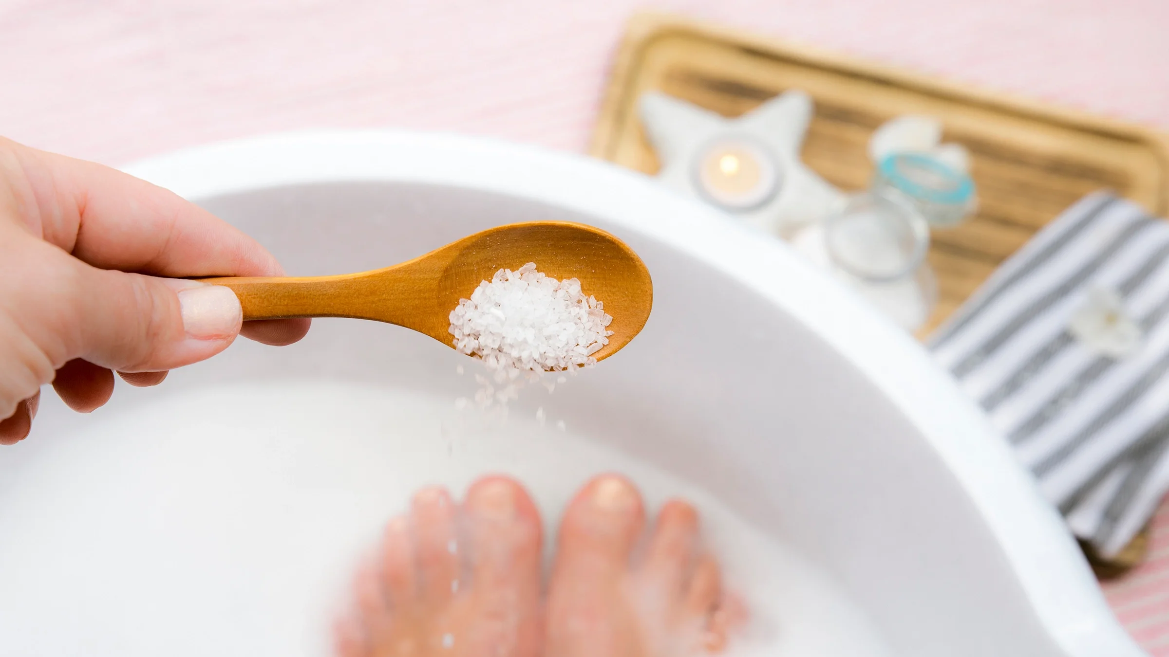 Close-up of person sprinkling epsom salts into foot bath from wooden spoon.