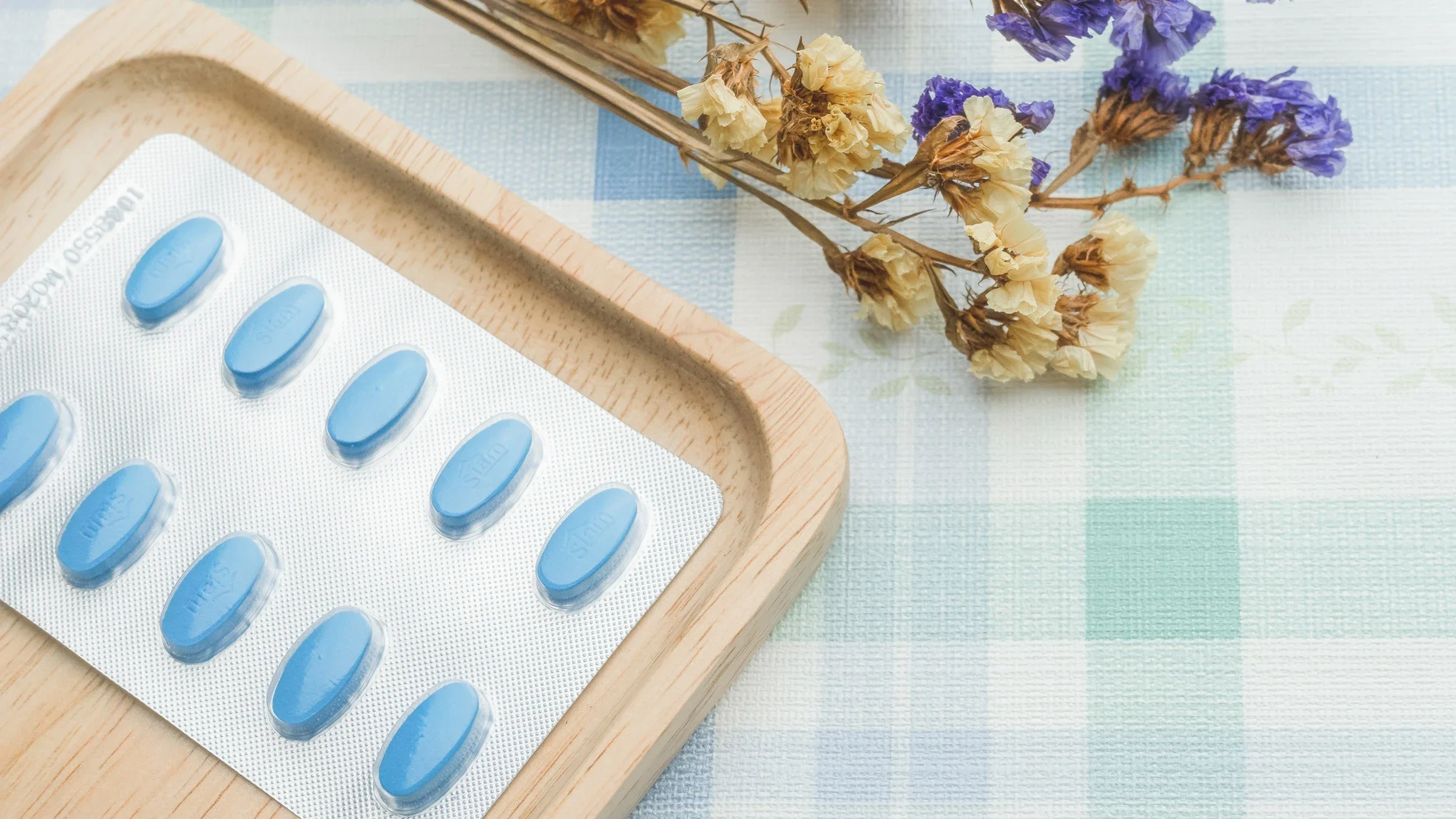 Blue pills on a wooden tray next to flowers on a plaid table cloth.
toons17/iStock via Getty Images 