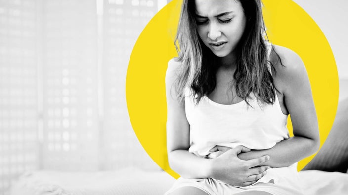 Black and white image of a young woman with long hair grasping her stomach in pain. She is sitting on the edge of her bed. There is also an added yellow graphic circle behind her head.