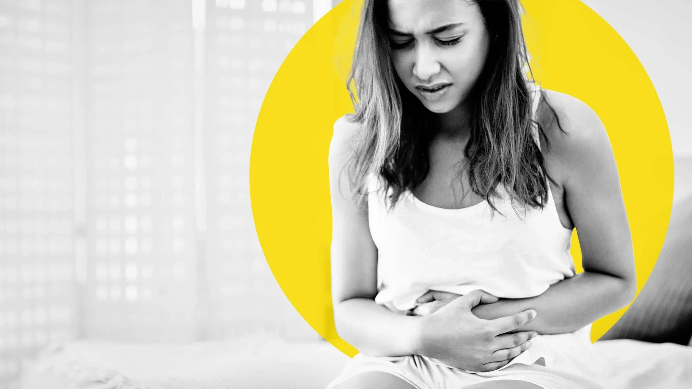 Black and white image of a young woman with long hair grasping her stomach in pain. She is sitting on the edge of her bed. There is also an added yellow graphic circle behind her head.