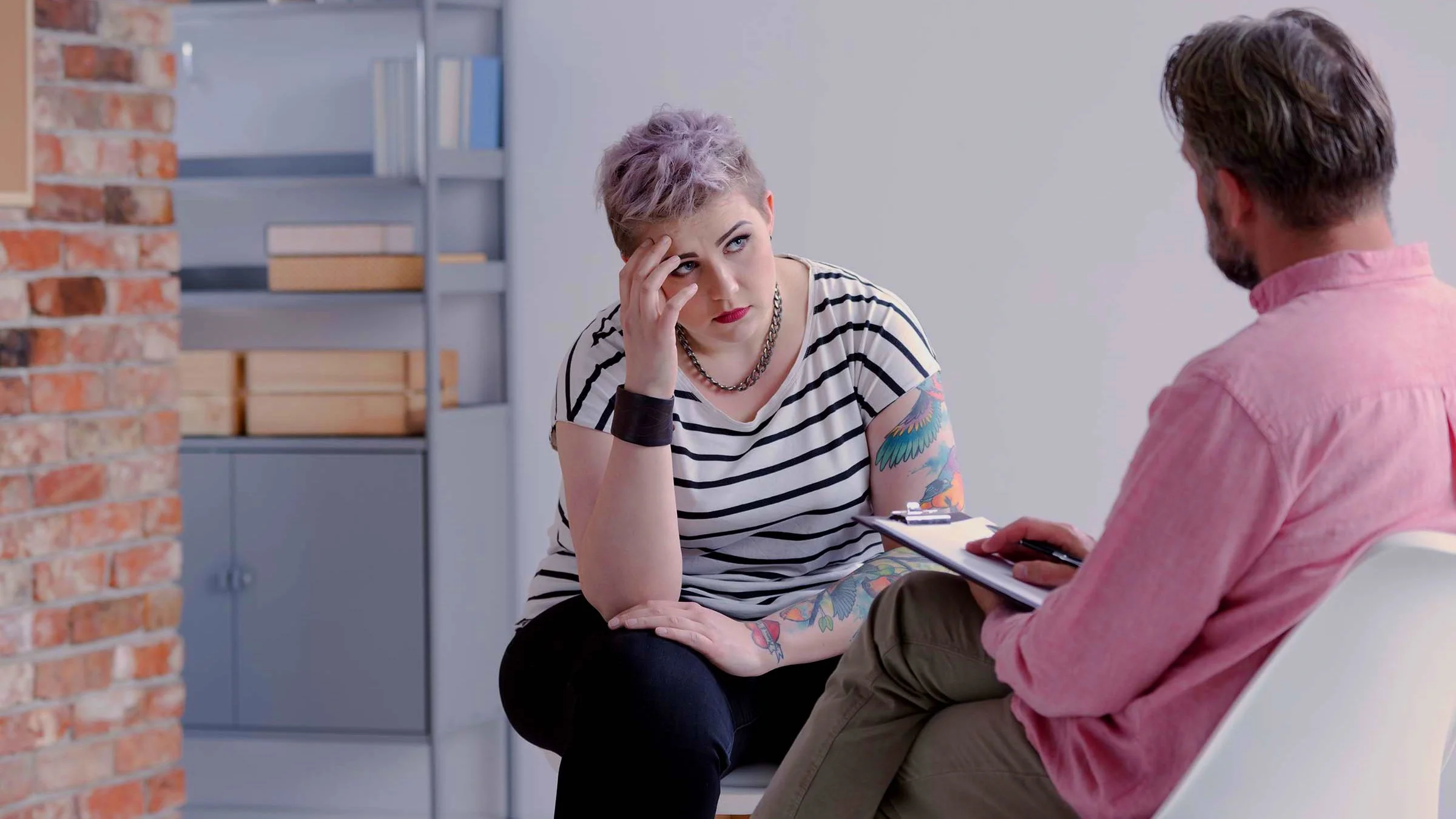 Young woman with short hair talking with her therapist. The therapist's back is to us while he talks to her and writes notes on his clipboard.