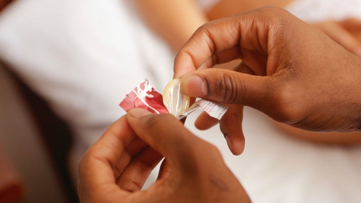 Close-up of a person opening a condom with white sheets in the background.