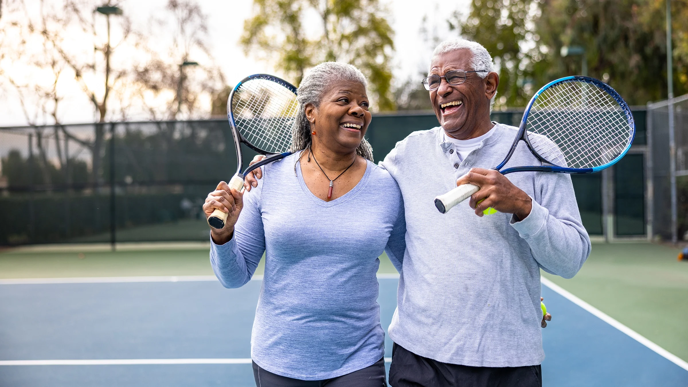 A couple laugh as they hold a pair of rackets at a tennis court. 