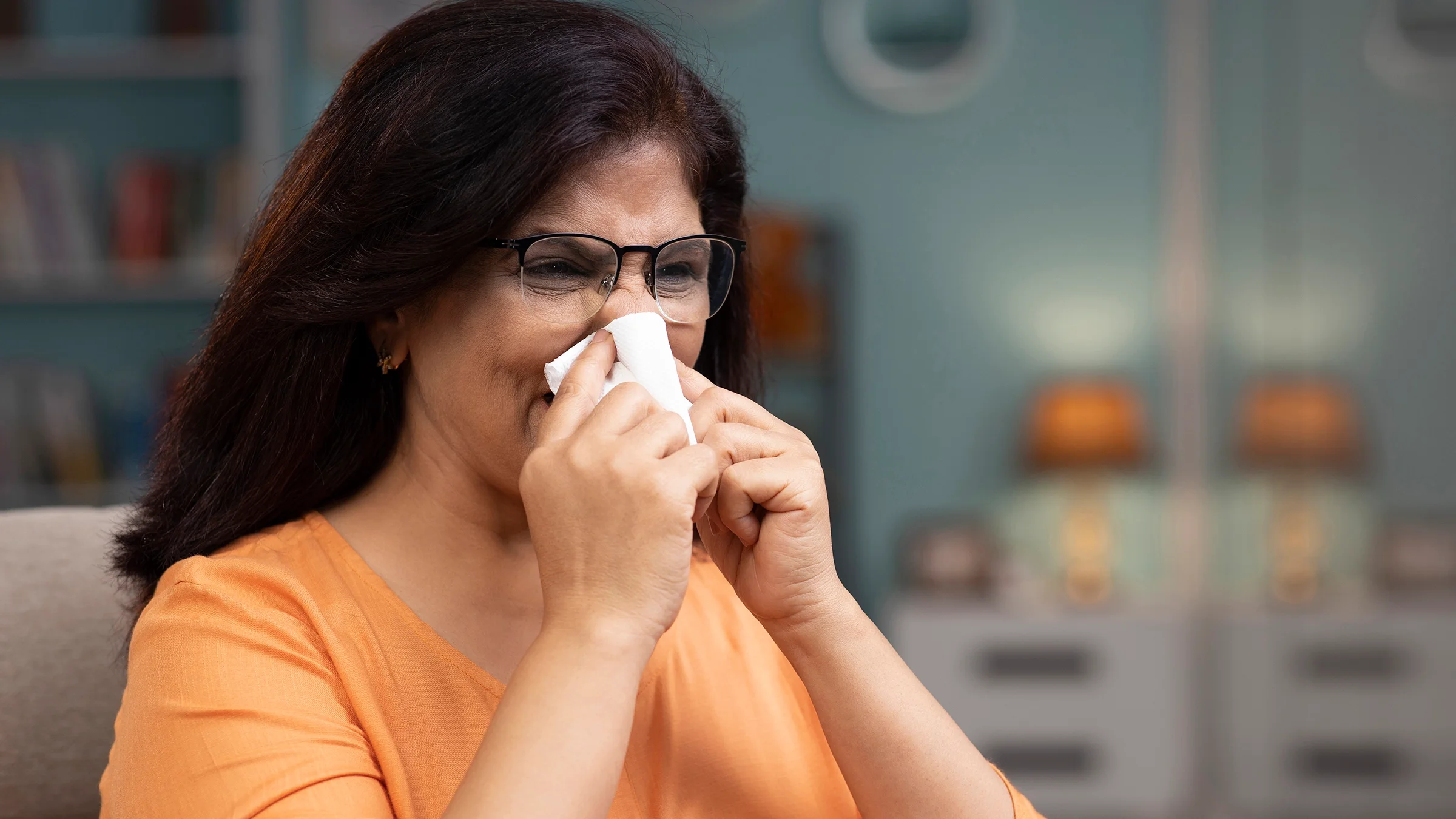 Woman with long hair and orange shirt sneezing into a tissue.