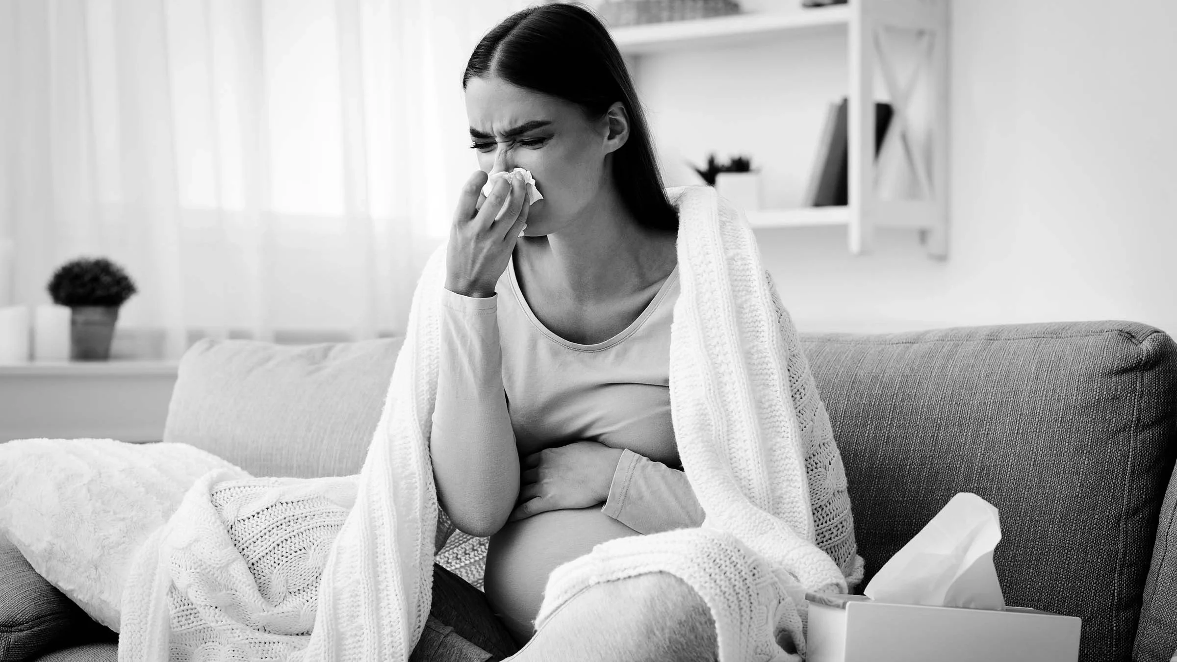 Black and white portrait of a pregnant woman blowing her nose. She is sitting on a couch wrapped in a blanket.