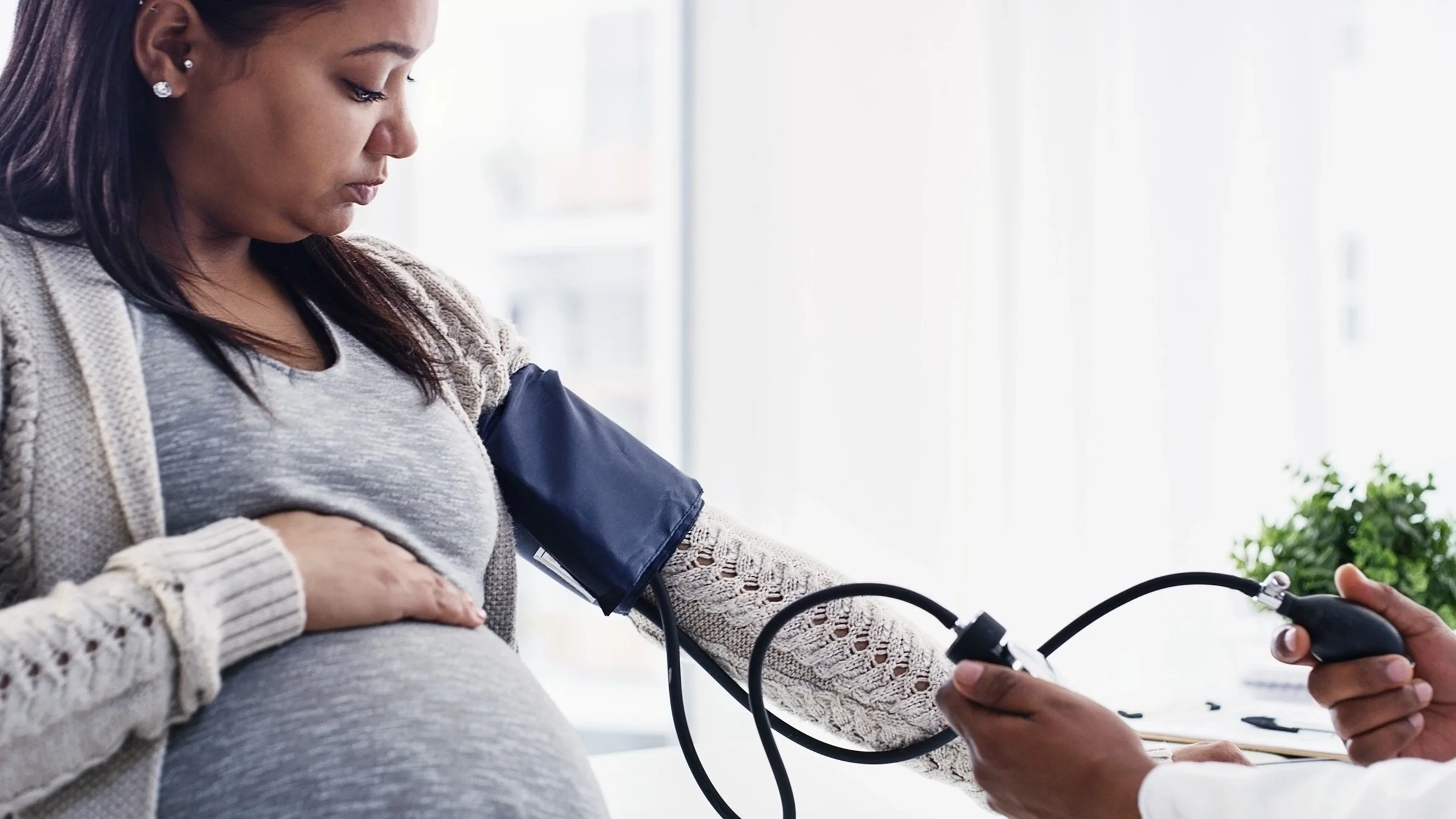 A doctor is checking a pregnant woman’s blood pressure. She has her left arm in the blood pressure cuff and is resting her right hand on her pregnant belly.