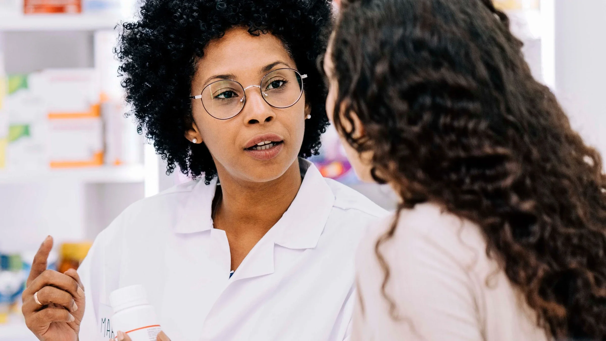 Young Black pharmacist talking to customer while holding a white medicine bottle.