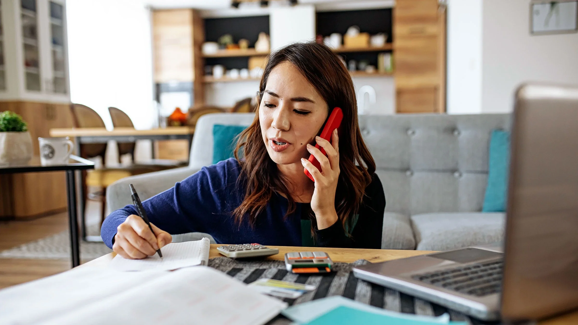 A woman is doing her finances at home, using her mobile phone and laptop.