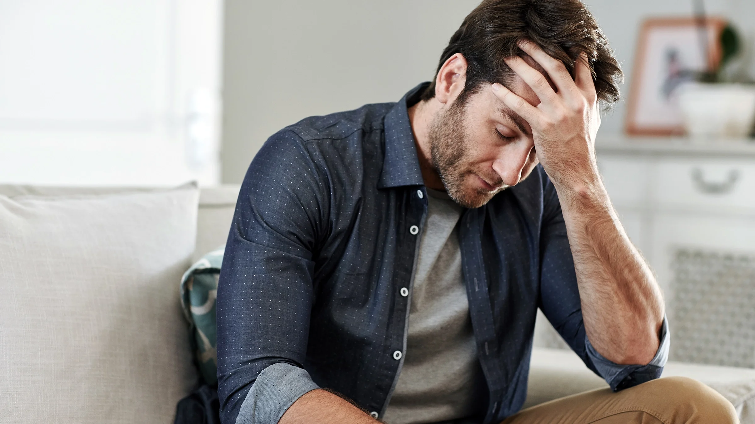 A man sits at home holding his head and looking distraught.