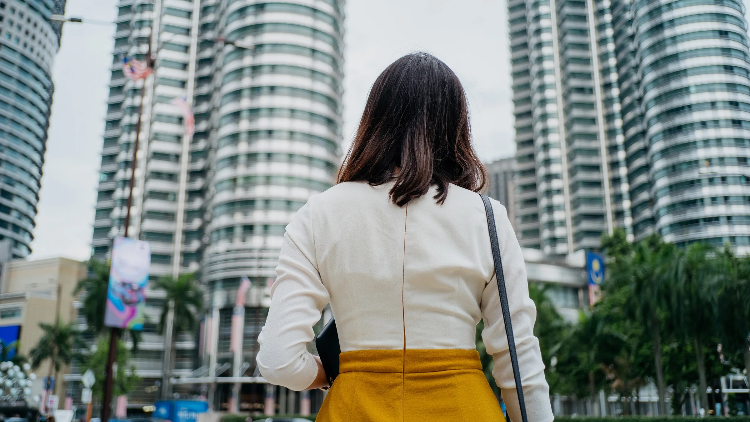 A woman is lost in a city with skyscrapers. The photo is of her back as she looks up at the buildings.