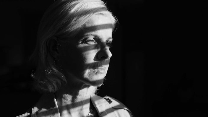 Black and white portrait of an older woman looking out a window with a shadow of lines across her face.
