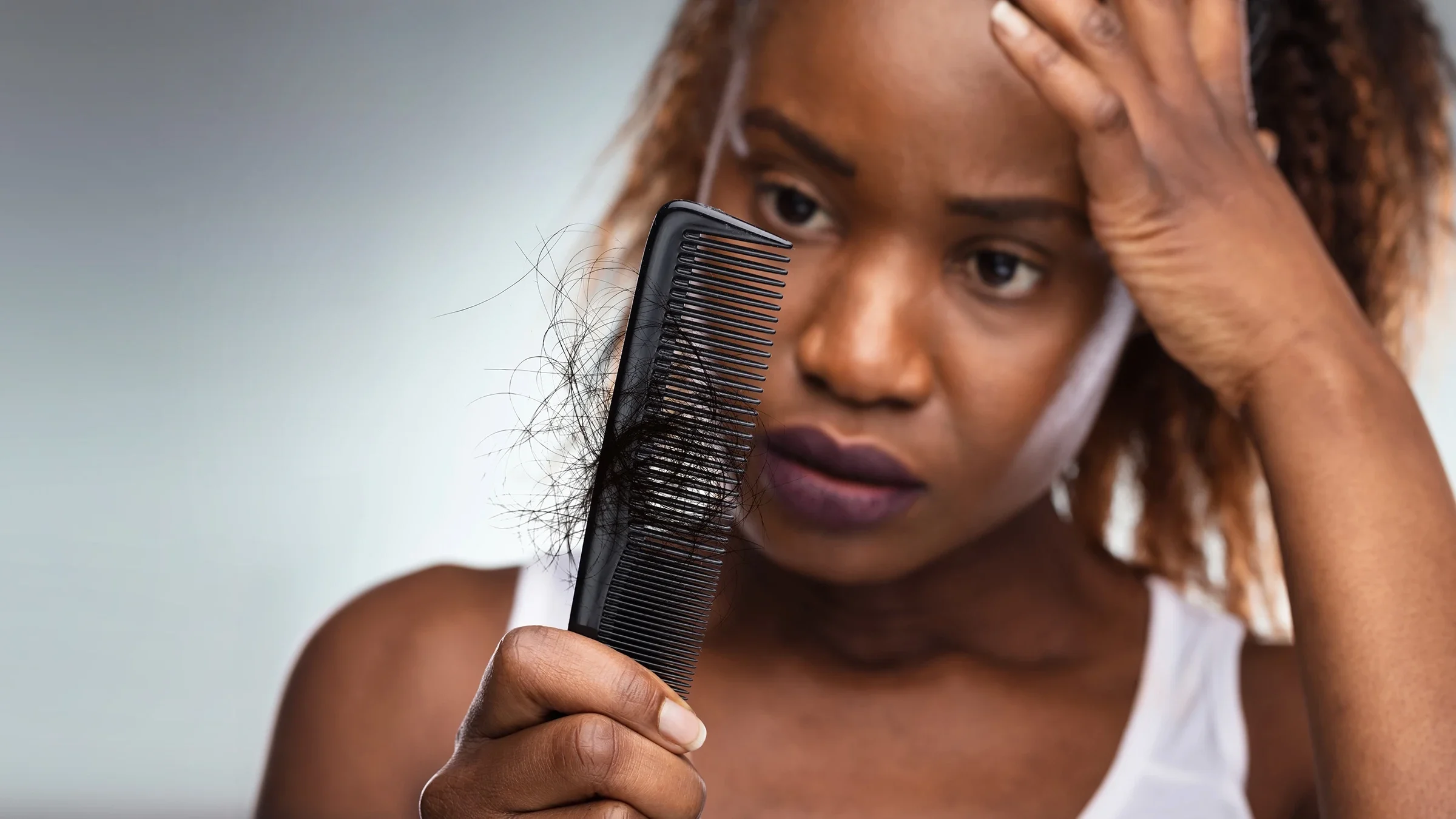 A woman clutches her forehead in distress as she examines a clump of hair in her comb.