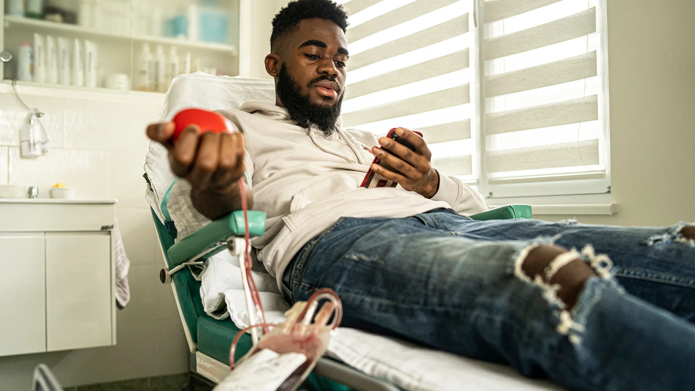 African American man donating blood at a clinic. He is squeezing a heart shaped stress ball while scrolling on his phone.
Vladimir Vladimirov/E+ via Getty Images