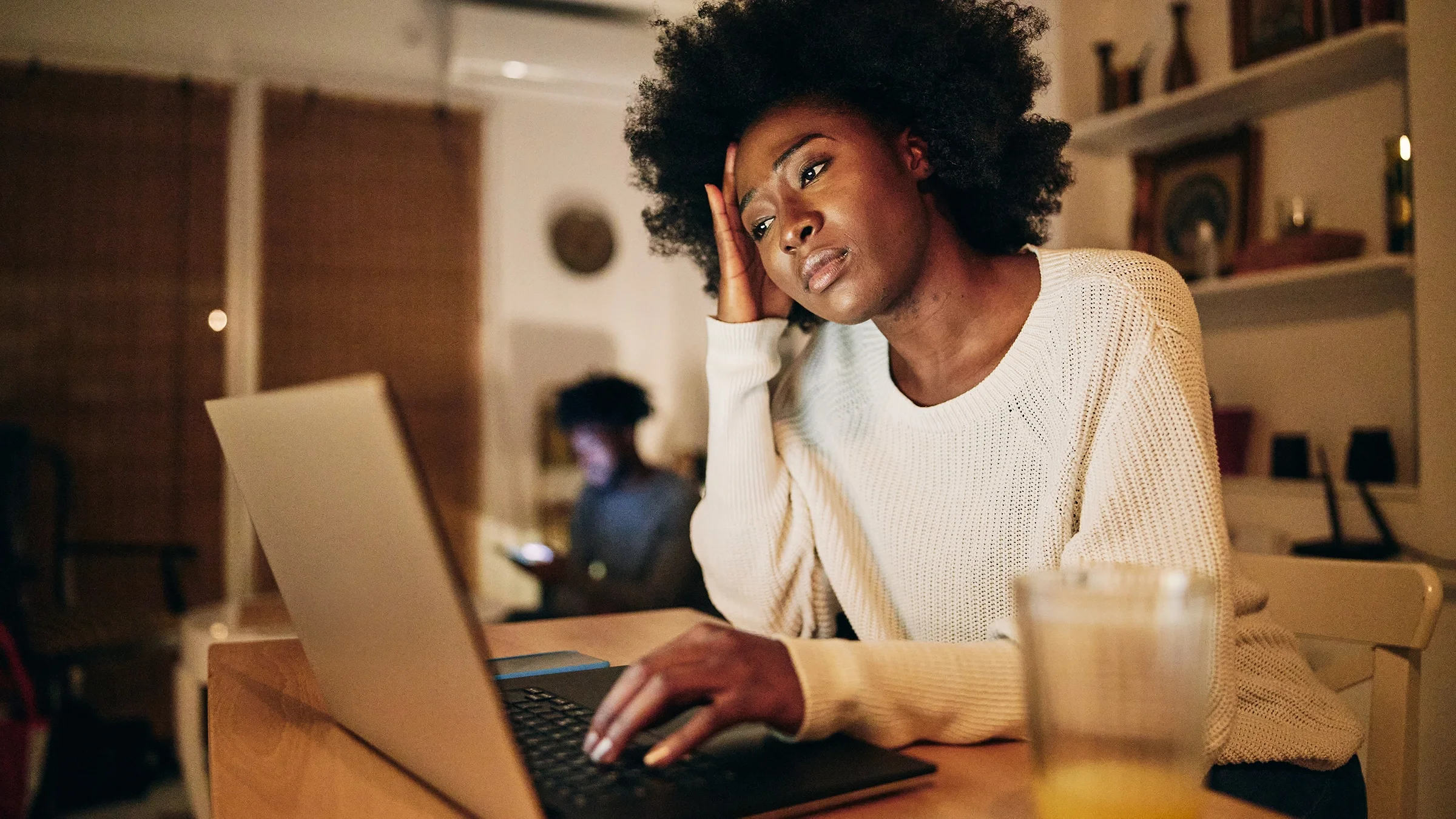 Young Black woman sitting at her laptop at the dining table with her hand on her head looking sad and frustrated.