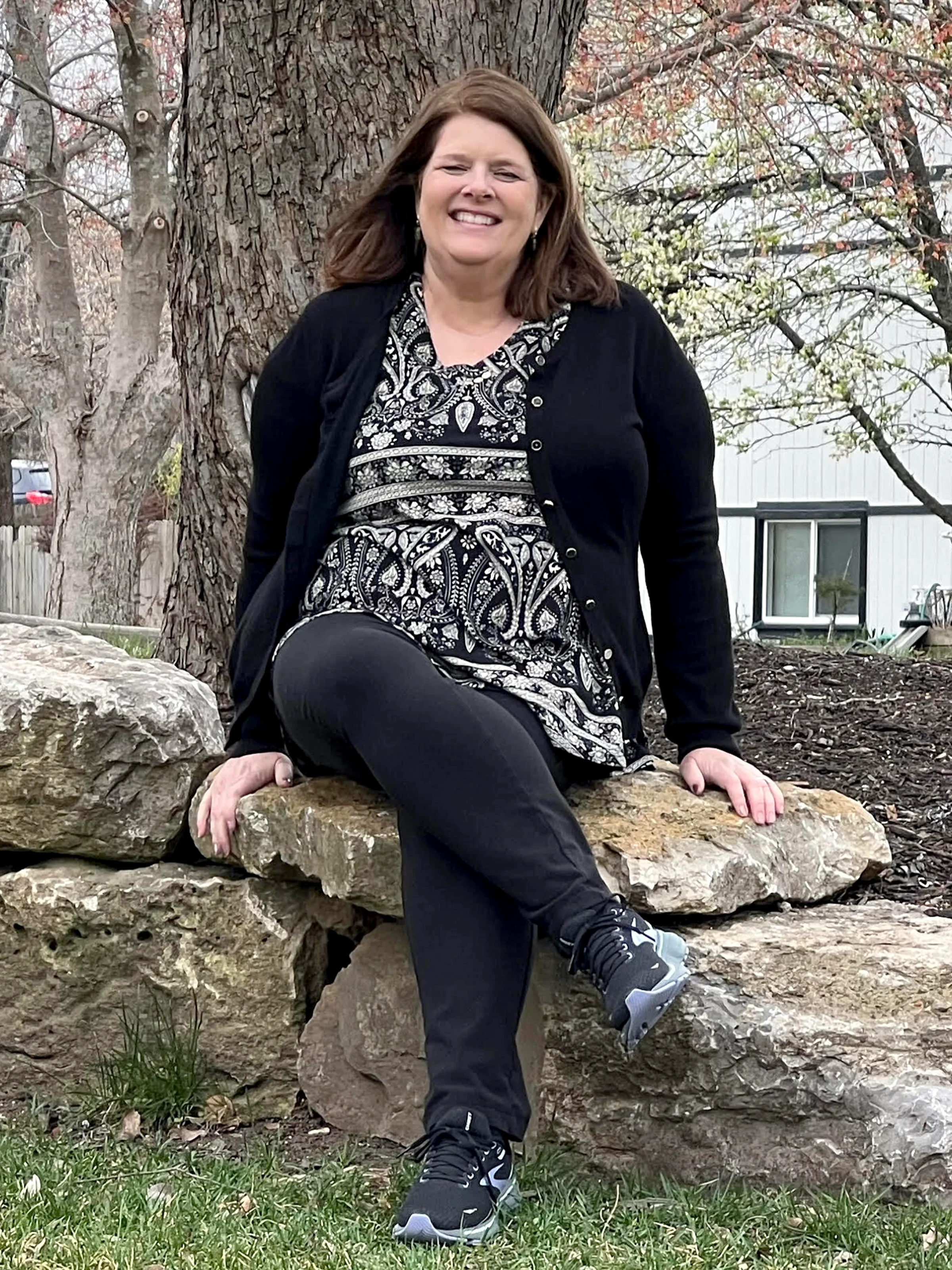 Kim Phillips is pictured outdoors, sitting on a rock wall in front of a tree.