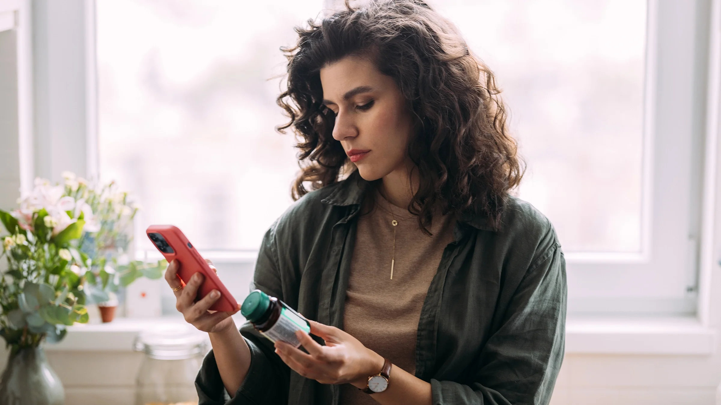 Woman holding a bottle of vitamins in one hand while looking at a smartphone.