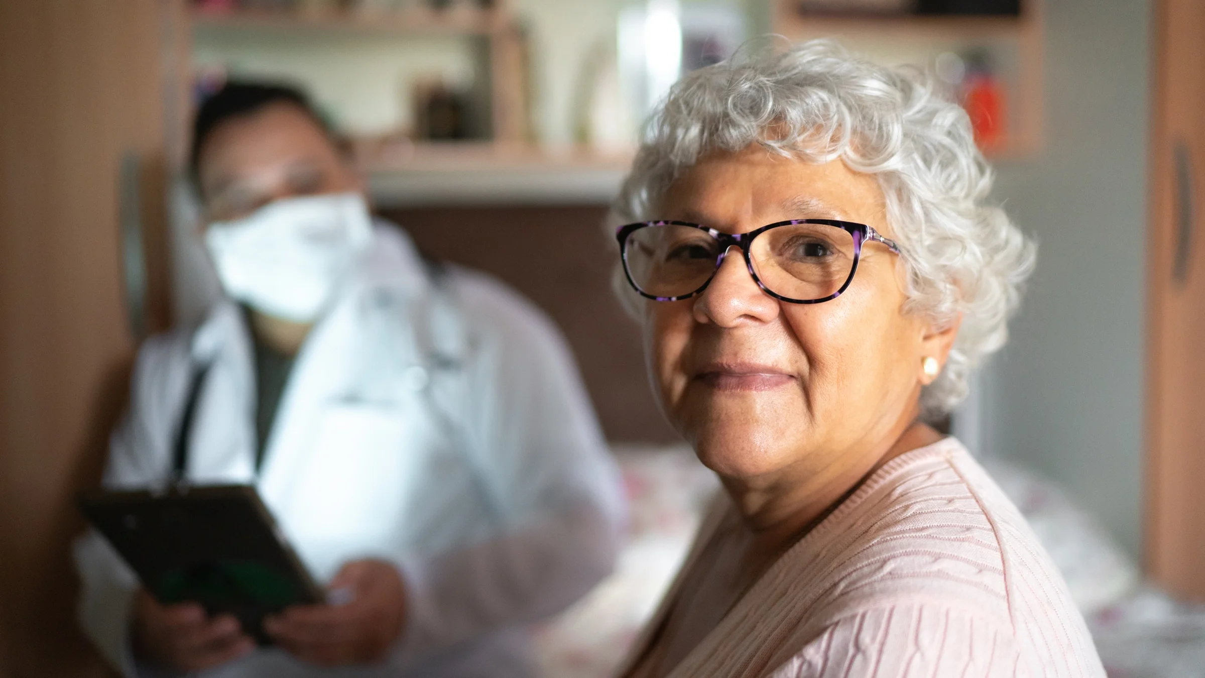 A senior woman looks into a camera with a healthcare professional in the background.