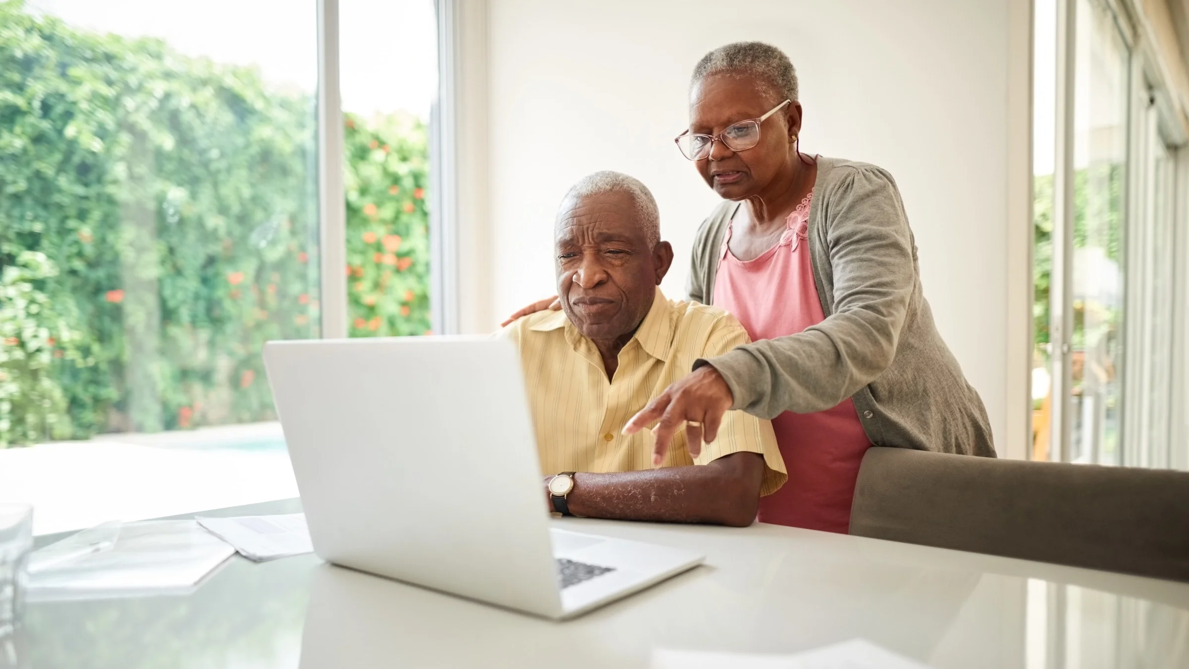 Elderly couple at dining table with laptop reviewing something on screen, the man is sitting while the woman stands behind him holding his arm.