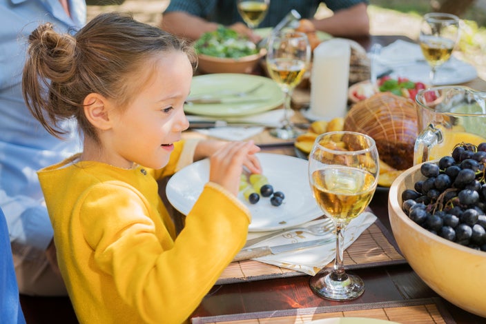 Young girl in a yellow sweater sitting at an outdoor dining table looking at a glass of white wine.