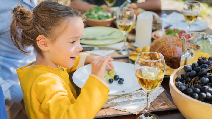 Young girl in a yellow sweater sitting at an outdoor dining table looking at a glass of white wine.