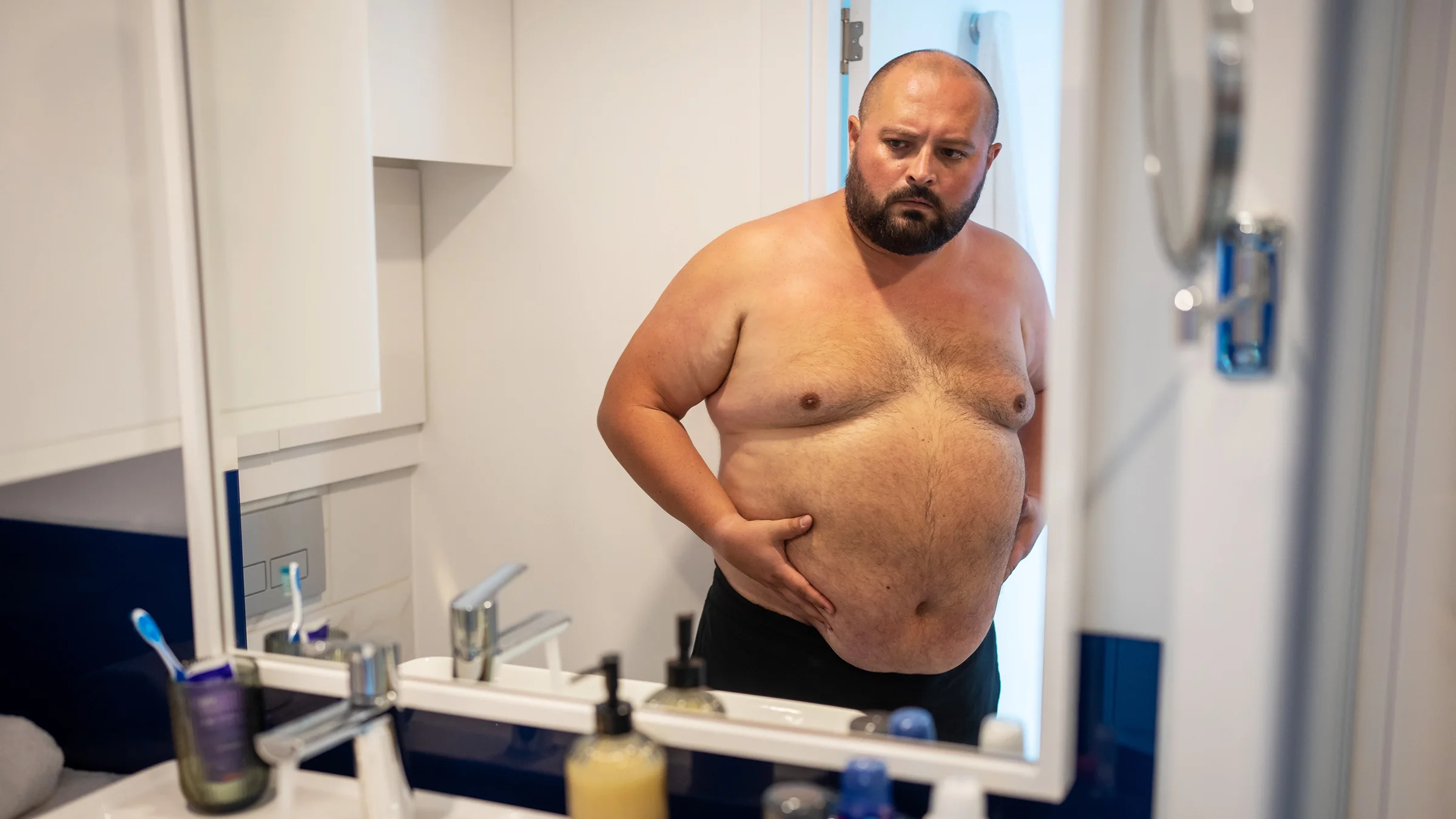 A shirtless man with excess breast tissue looks at himself in a bathroom mirror.