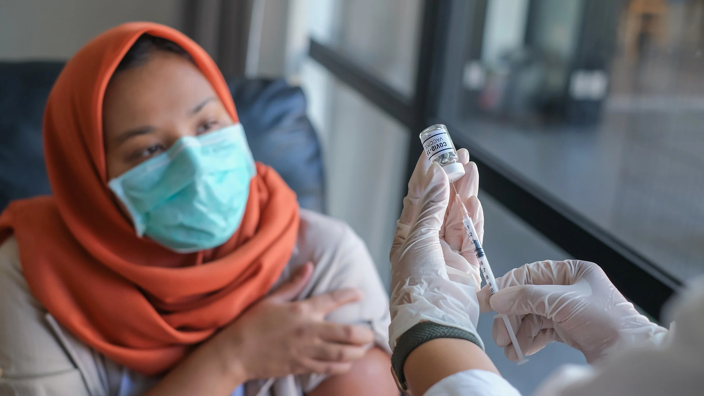Young woman with a headscarf on rolling up her sleeve as a nurse prepares the COVID-19 vaccine.