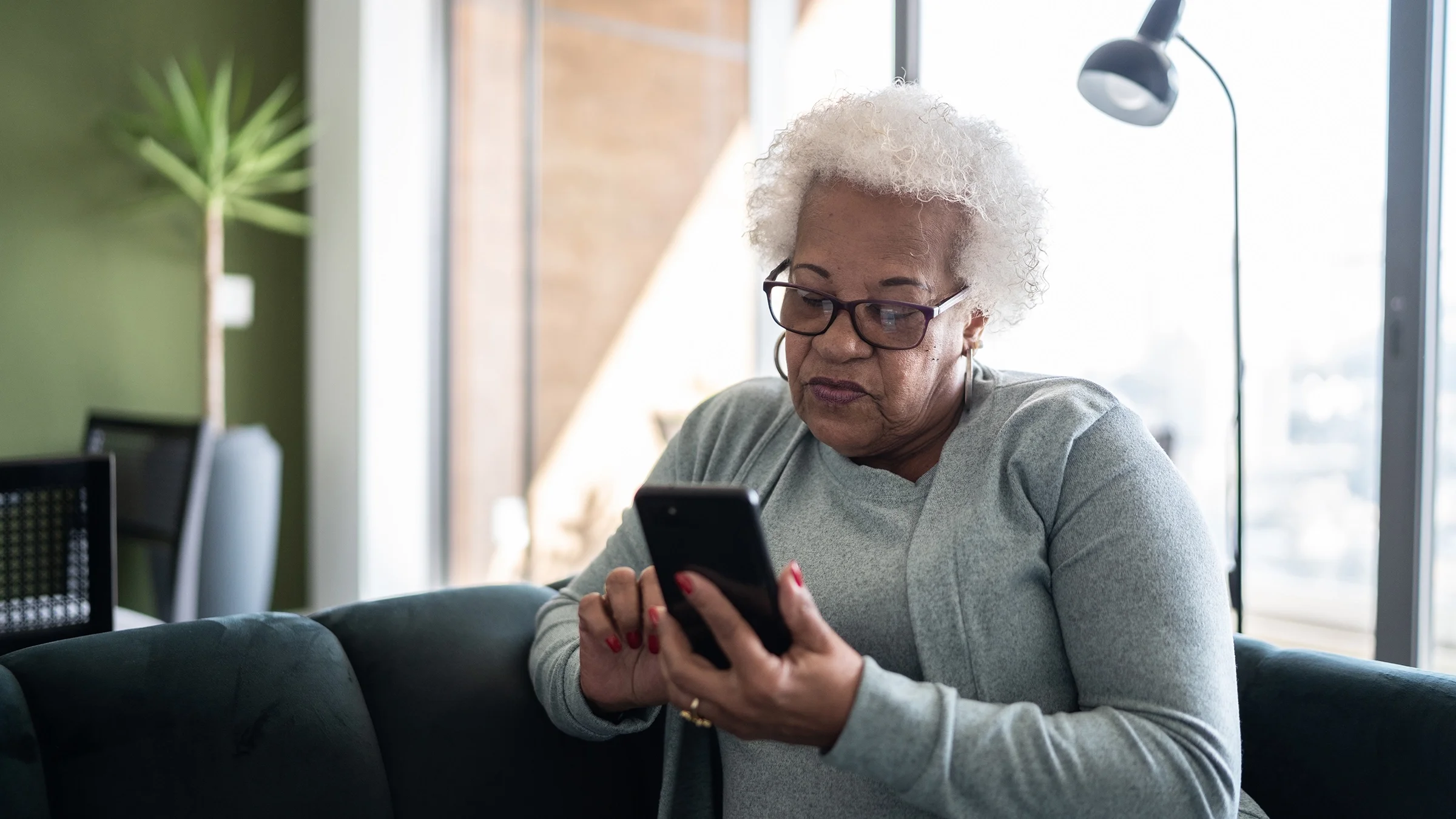 Senior woman with short white curly hair. She is sitting on the couch using her smartphone.
