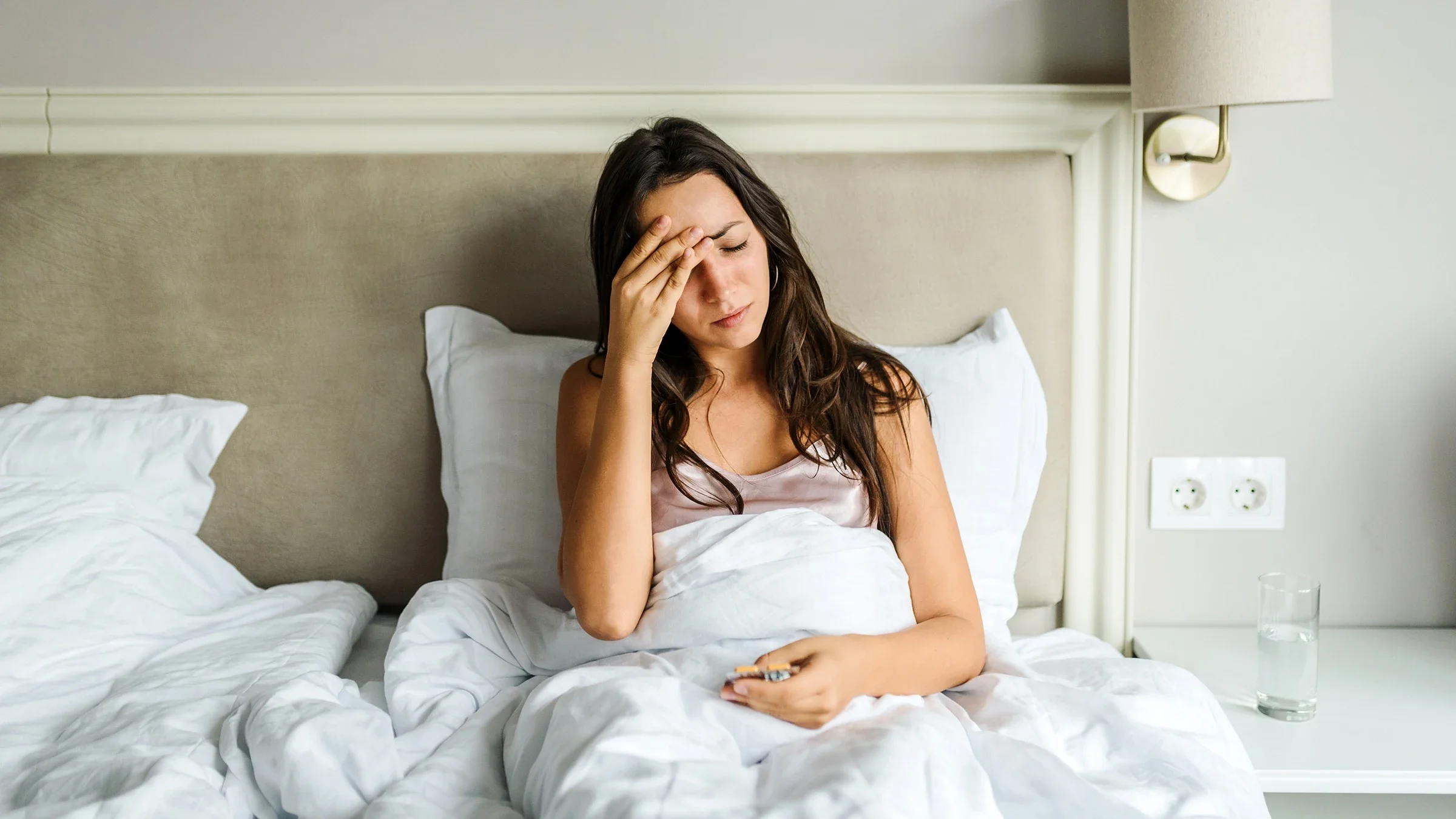 Young woman sitting up in bed with her hand on her forehead in pain.
Vasil Dimitrov/E+ via Getty Image